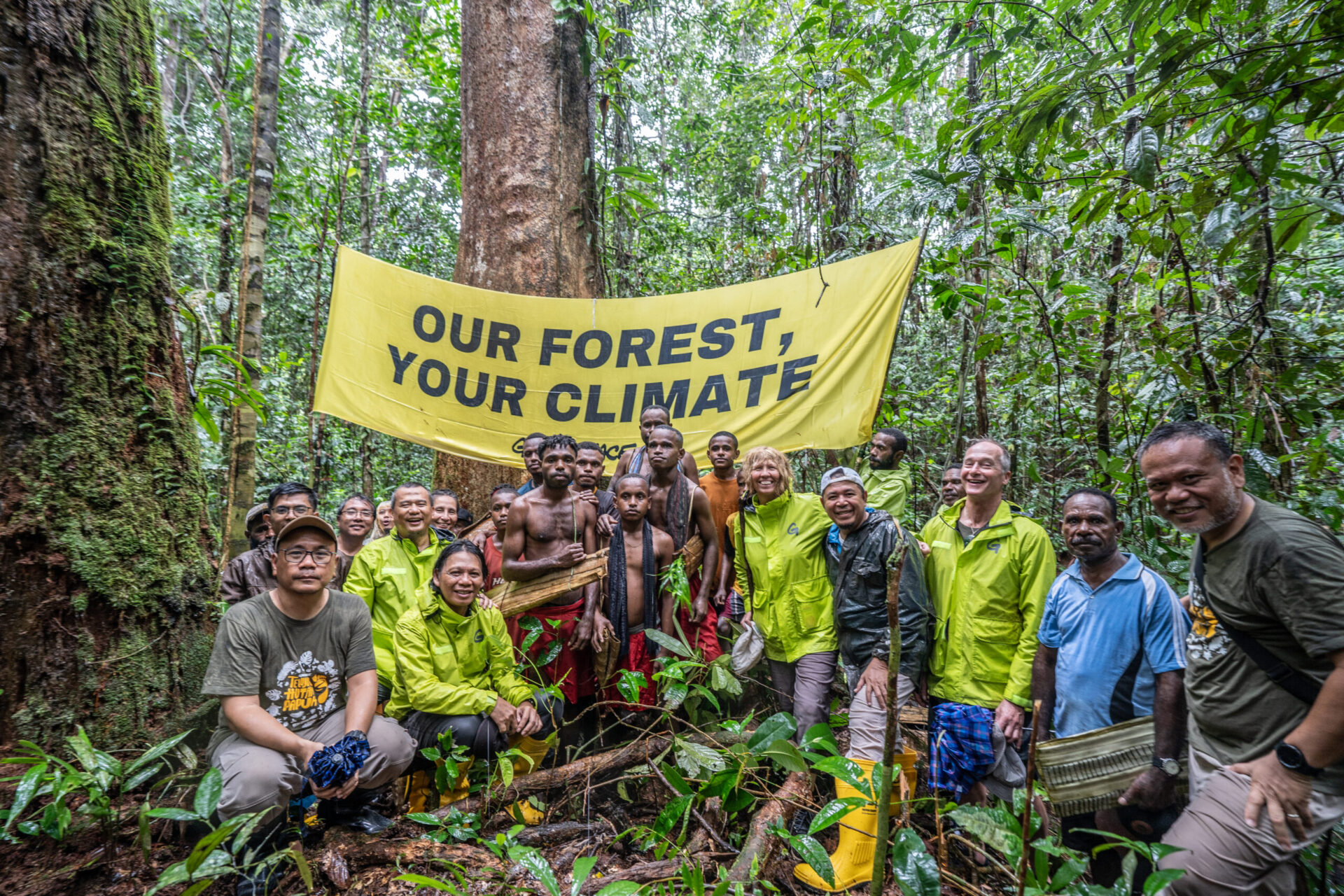 A crowd of around 20 people crowd in front of a banner reading Our Forest Your Climate, inside a lush rainforest
