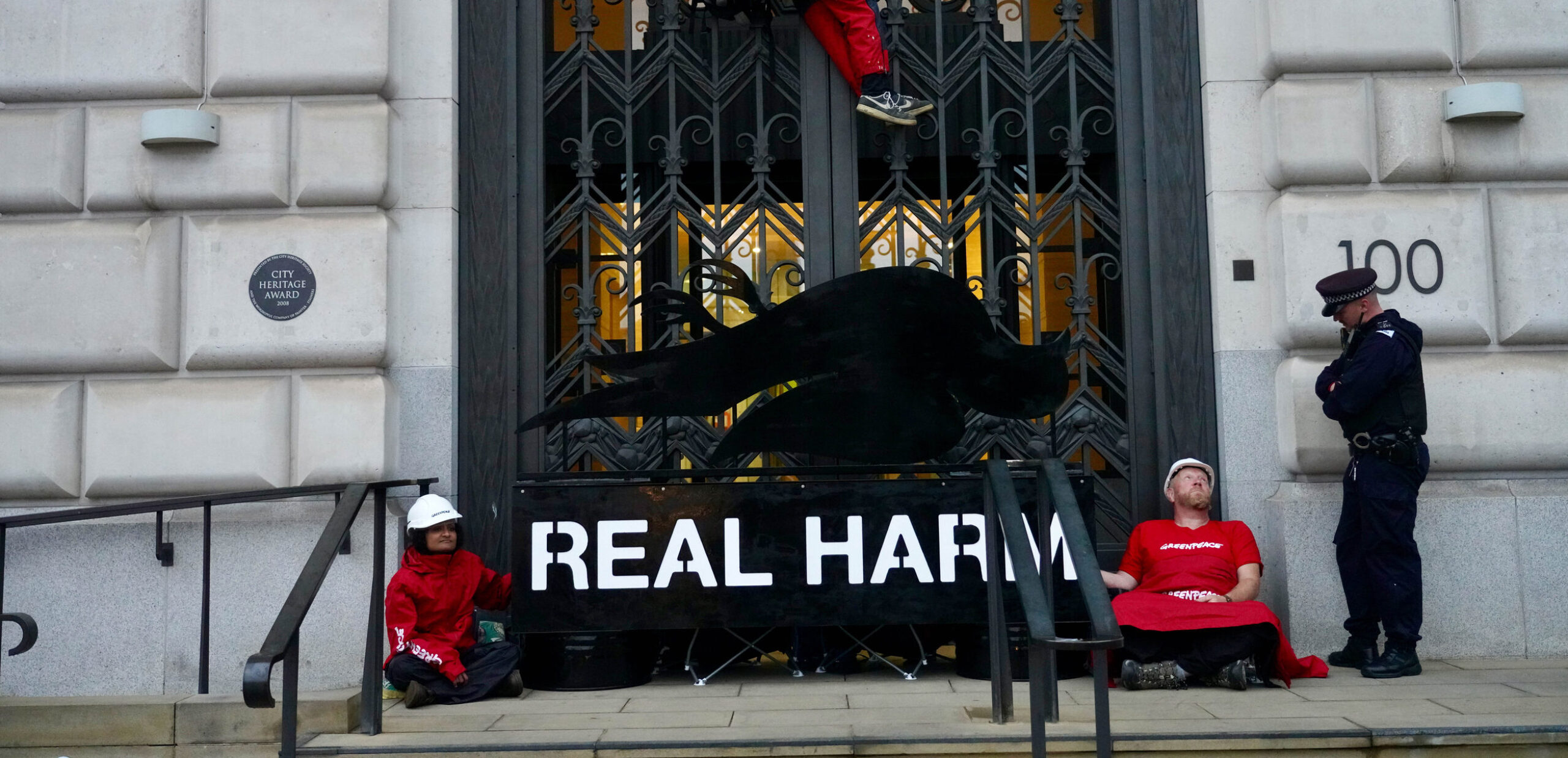 Two activists are locked on to a subverted Dove branded logo with an image of a dead dove, blockading the entrance to a doorway, while a climber is suspended above them and one policeman speaks to one of the activists