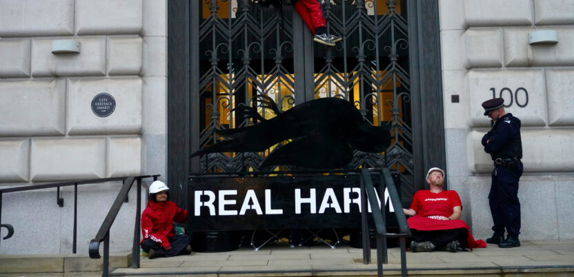 Two activists are locked on to a subverted Dove branded logo with an image of a dead dove, blockading the entrance to a doorway, while a climber is suspended above them and one policeman speaks to one of the activists