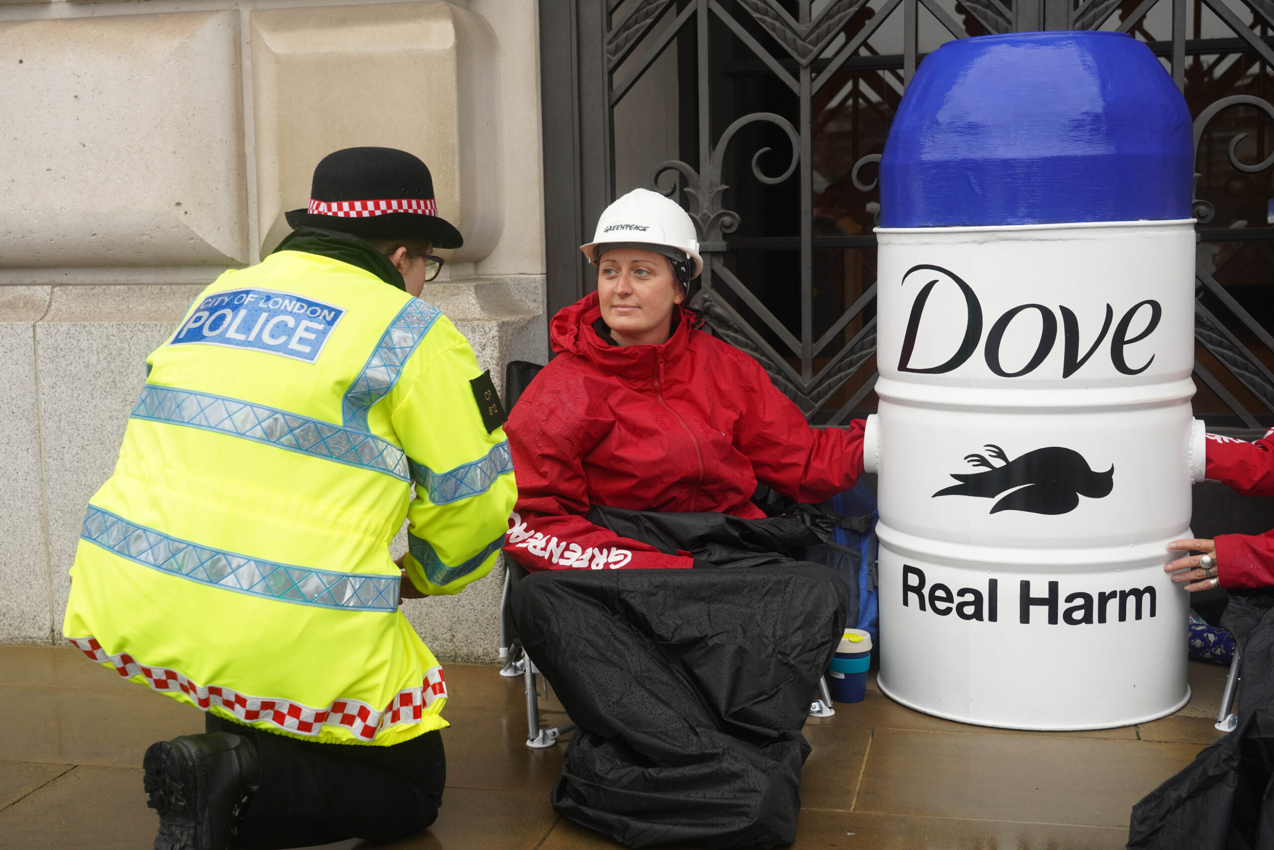 An activist locked to a barrel adapted to look like a Dove deodorant container blocks the door of a grand building. A police officer is crouching down to speak to her.