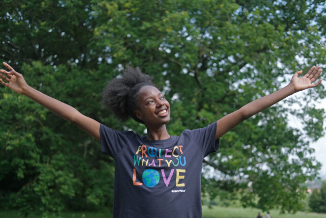 In a lush green woodland, a young woman in a 'protect what you love t-shirt' spreads her arms wide and smiles.