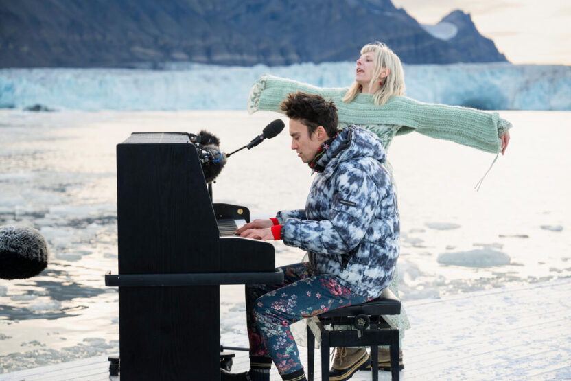 Jacob Collier is sat at an upright piano next to Aurora singing on a floating platform in the sea with a glacier in the background