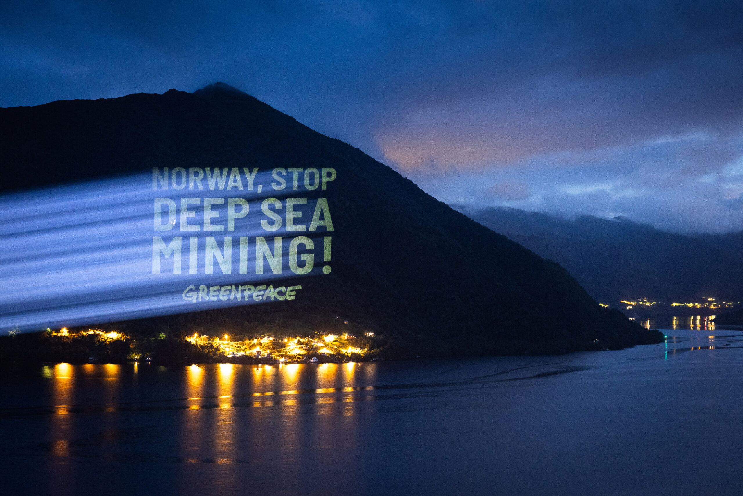 The words Norway, Stop Deep Sea Mining! Greenpeace are projected on a rock in the evening. Lights can be seen in the foreground.