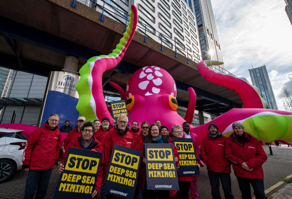 Greenpeace activists holding "Stop Deep Sea Mining" placards standing in front of a gigantic, pink octopus outside the Hilton hotel.