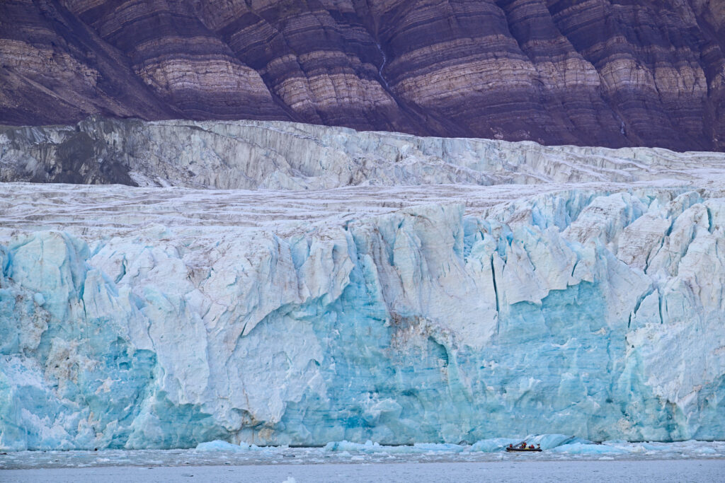 A small inflatable boat travels in front of an enormous iceberg in the Arctic.