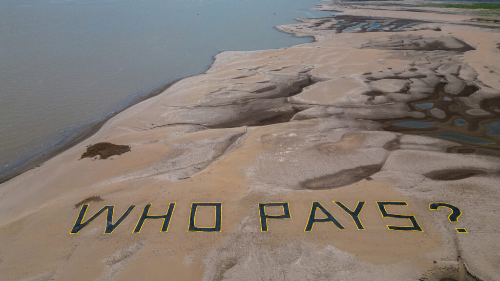 A dried out riverbed is seen from above with a banner saying 'Who Pays?'