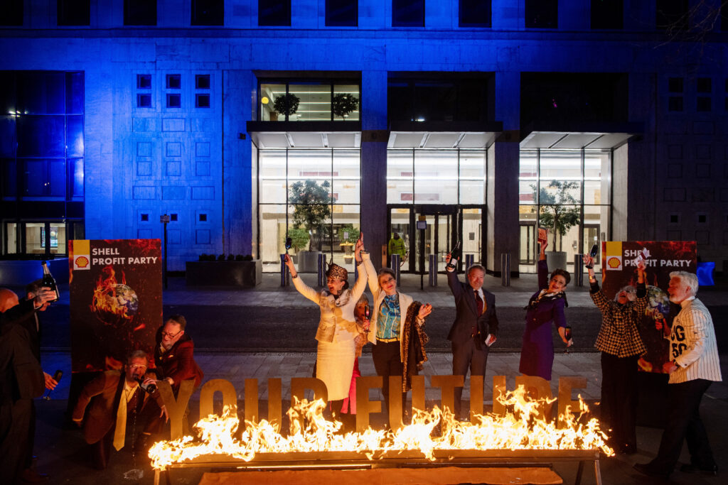 A sign saying Your Future is burning in the foreground with Greenpeace activists dressed as executives dancing and cheering behind it.
