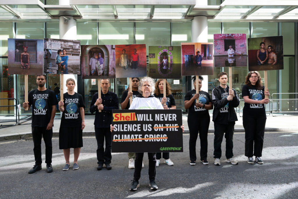 Activists hold up placards with images of climate impacted people outside the court. The activist in the foreground holds a sign which says 'Shell will never silence us on the climate crisis'.