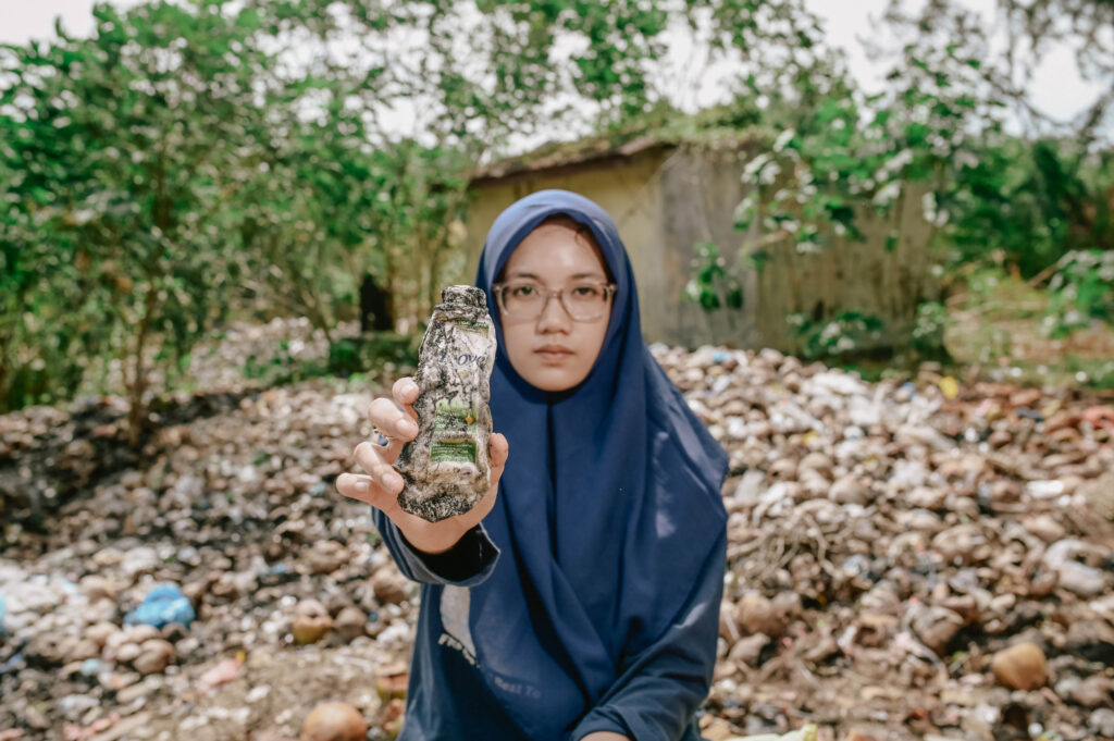 An activist sits in front of a plastic rubbish dump and holds a plastic Dove bottle into the camera.
