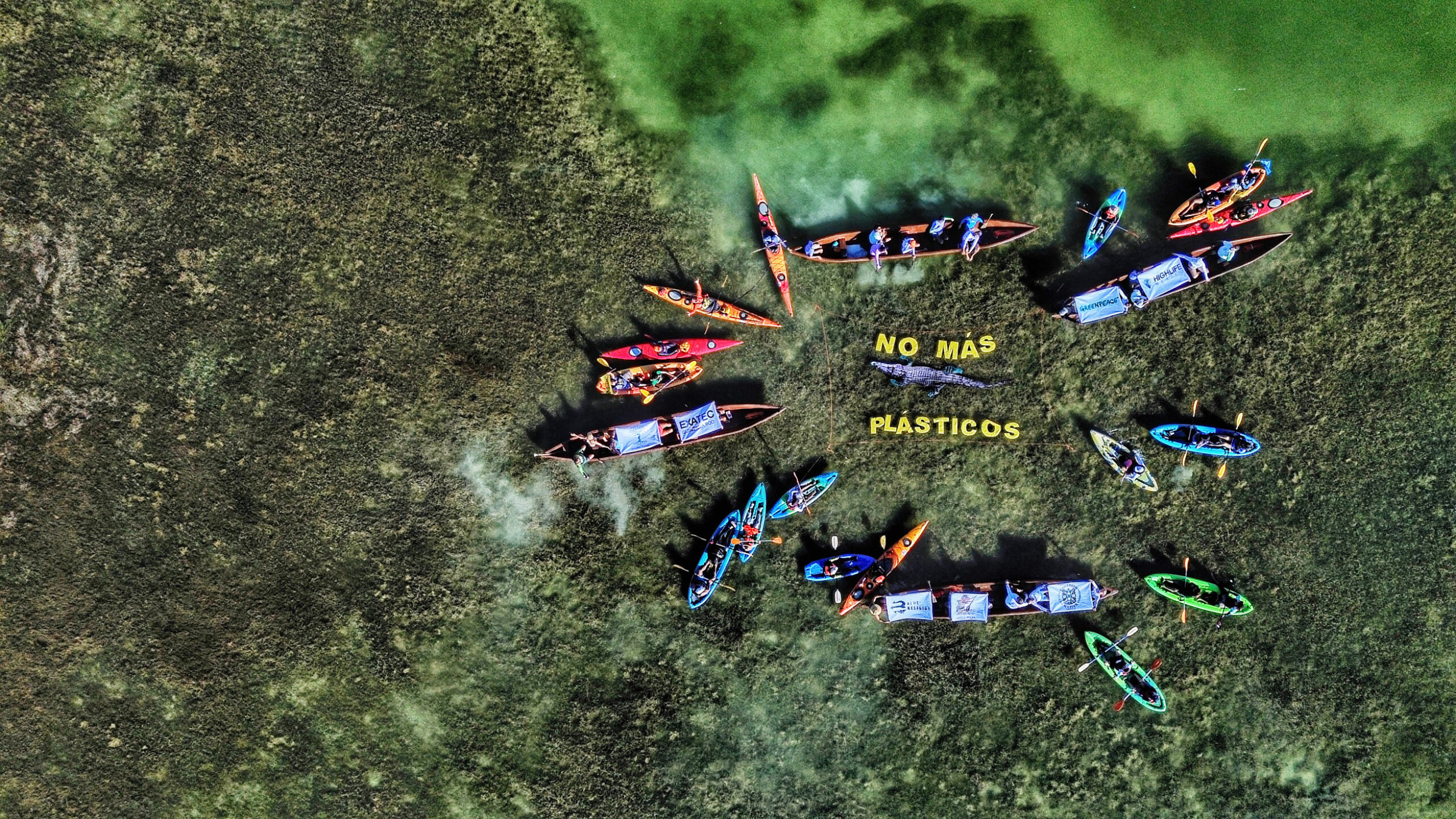 Aerial view of boats in a green lagoon with a banner saying 'No Mas Plasticos', meaning "no more plastics", in the middle