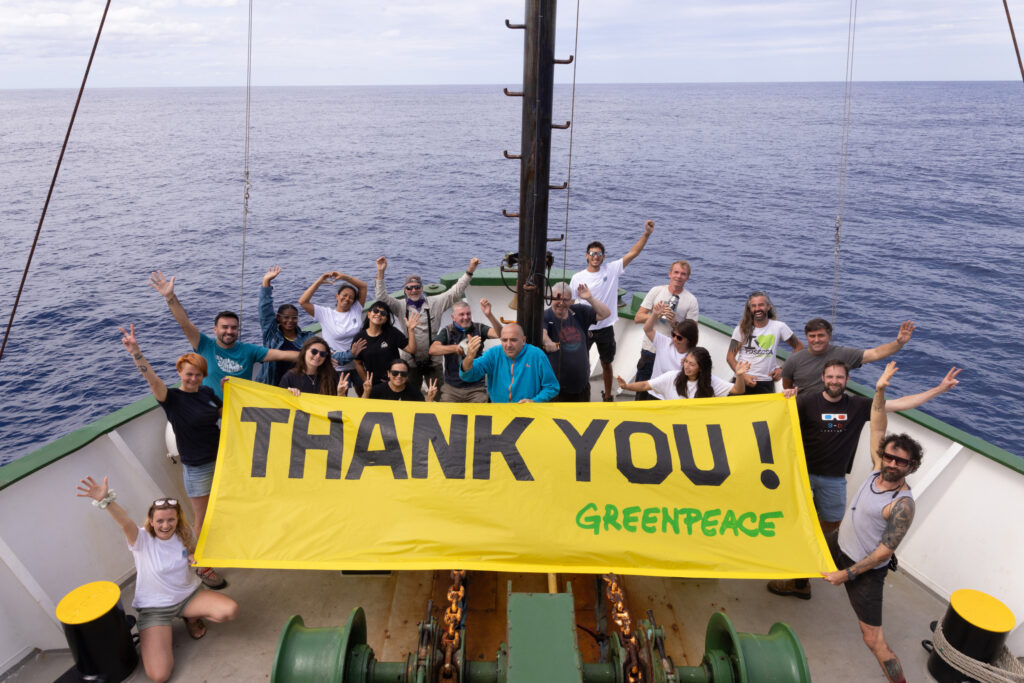 Group shot of campaigners on the Arctic Sunrise with a yellow Thank You banner with the ocean in the background.