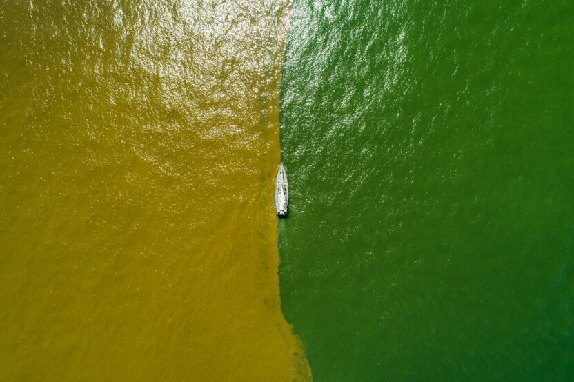 Aerial view of a boat sailing along a line where the brown water of a river and the green water of the ocean meet.