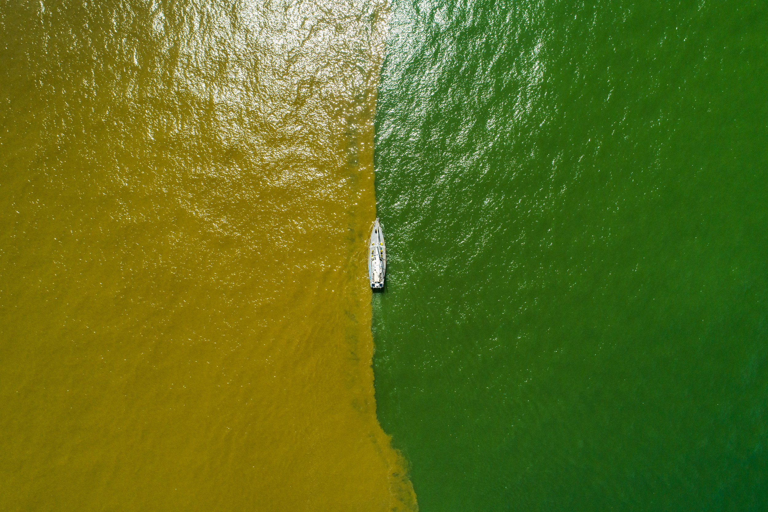 Aerial view of a boat sailing along a line where the brown water of a river and the green water of the ocean meet.