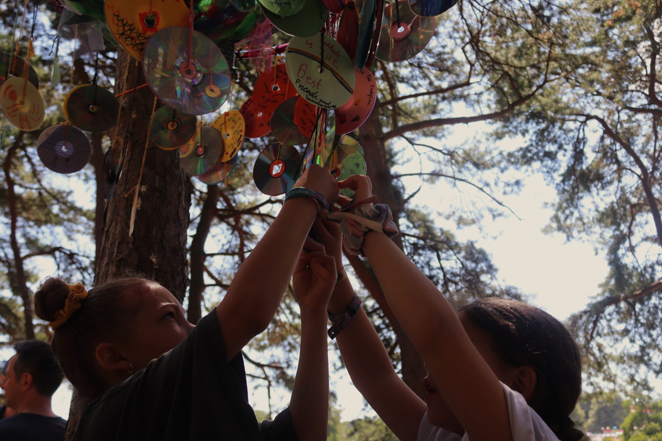 Two children reach above their heads to tie a personal message written on a compact disc to the branch of a tree with string.