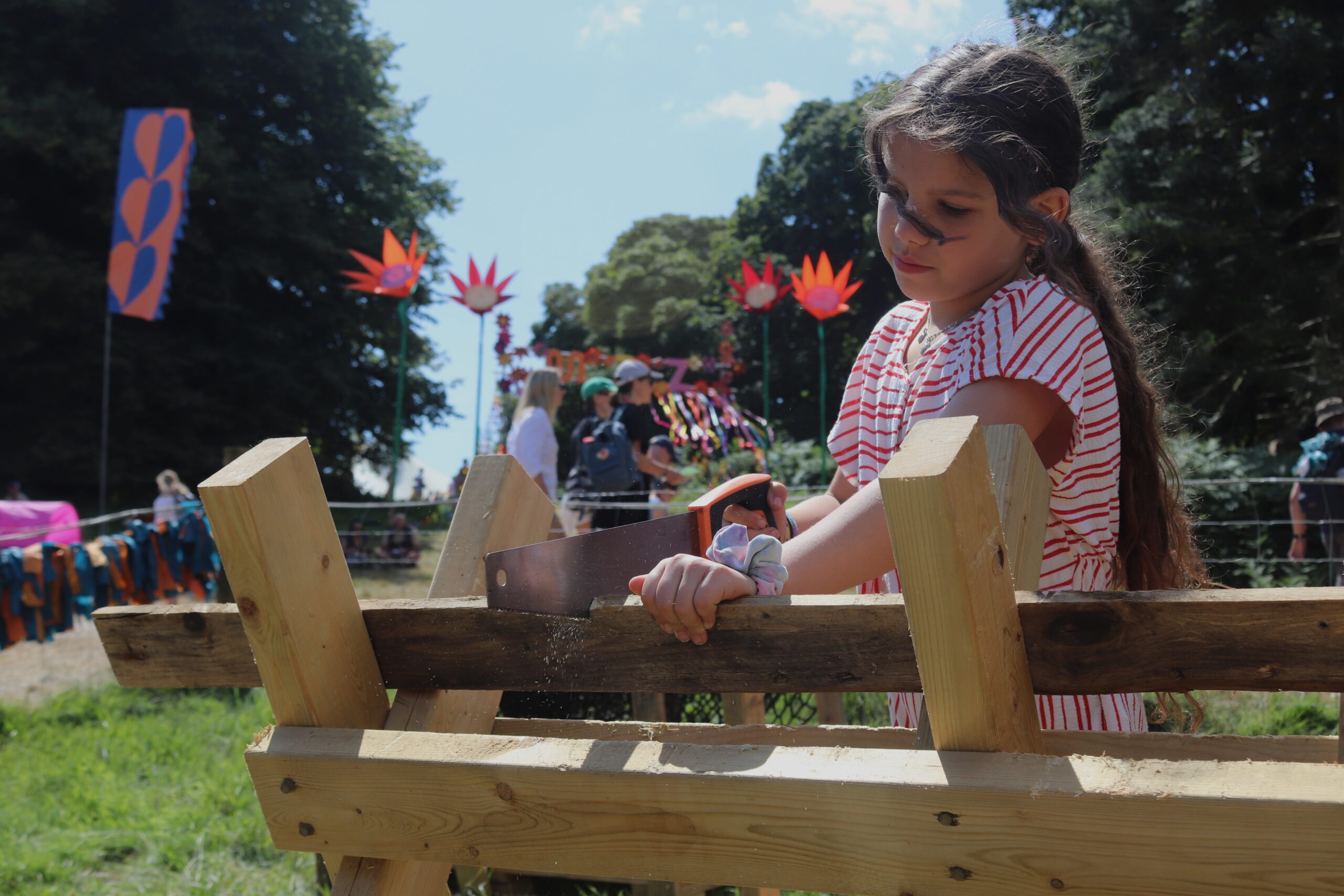 A girl uses a saw to cut a piece of wood using a wooden stand outdoors at Latitude