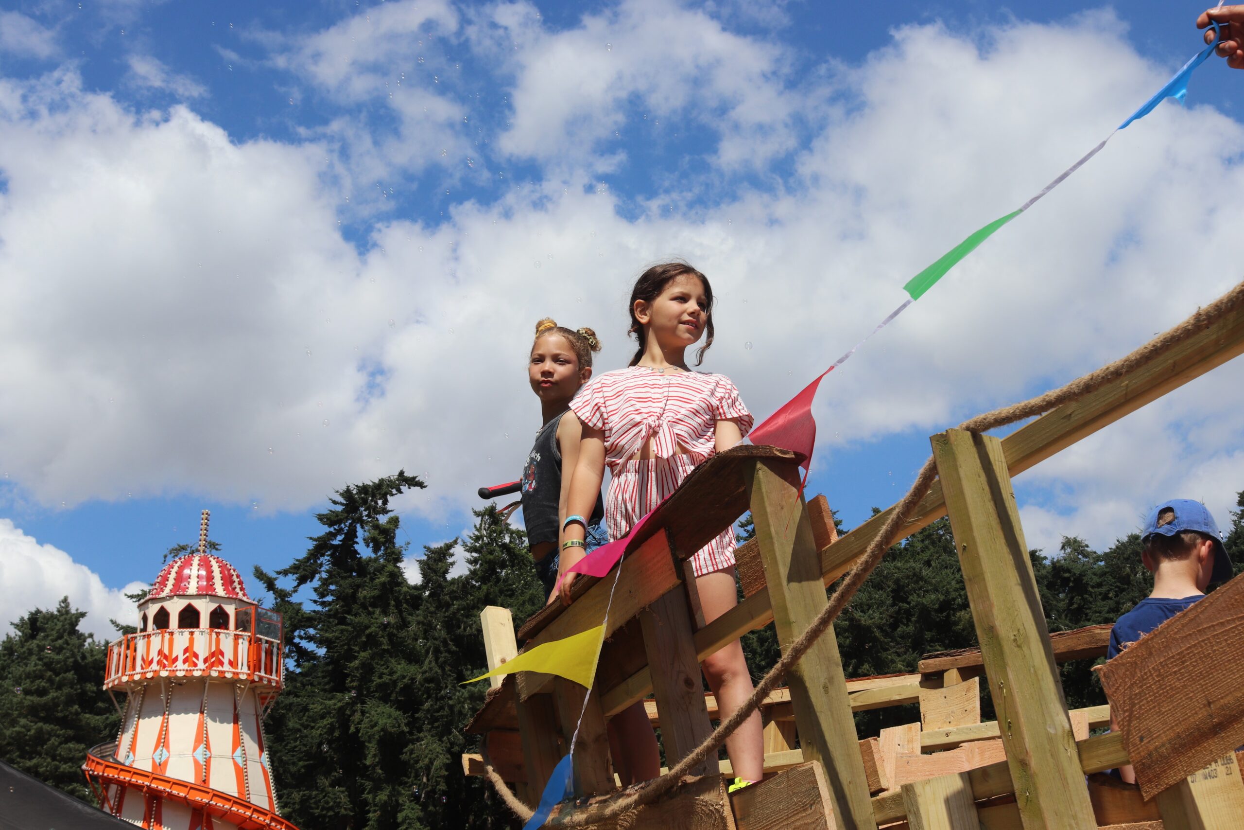 Two girls stand on top of a wooden climbing frame with a string of bunting and a helter skelter in the background.