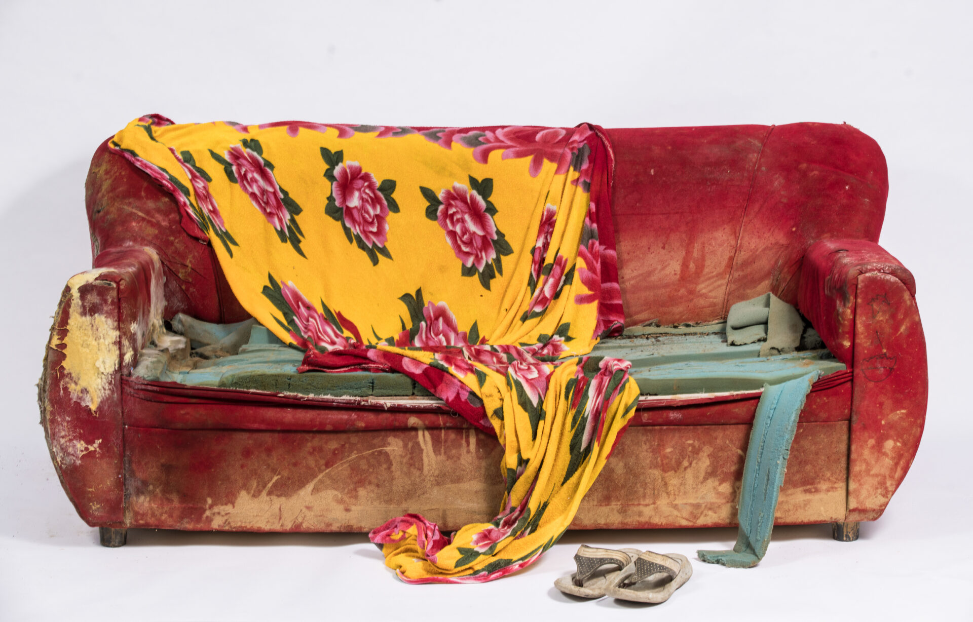 Broken red sofa with a yellow blanket with red roses draped over it with grey flipflops placed on the floor in front of it.