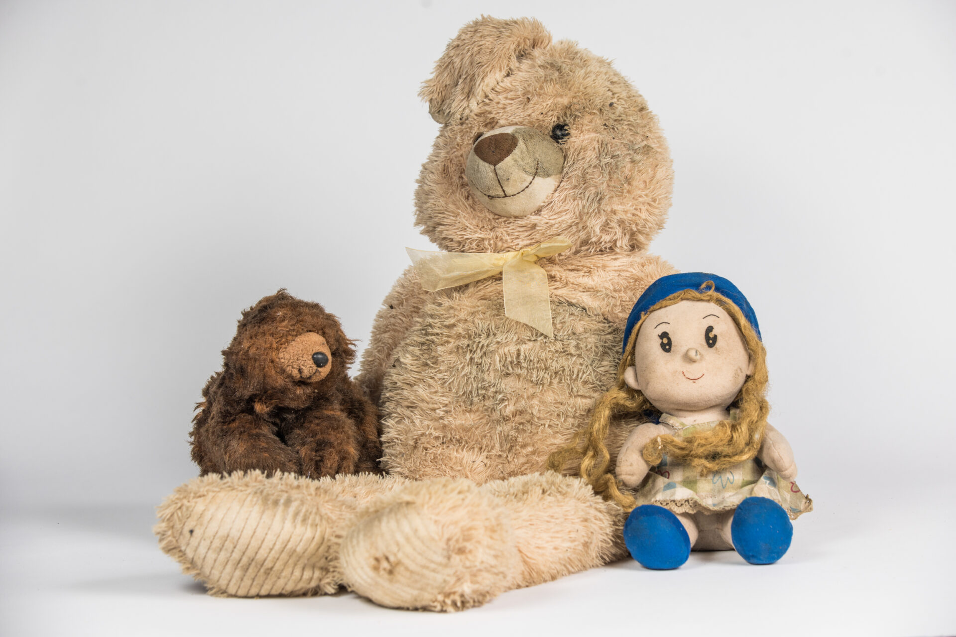 Various toys are photographed against a white background. A rabbit bear sits in the middle with a doll to its right and a brown bear to the left
