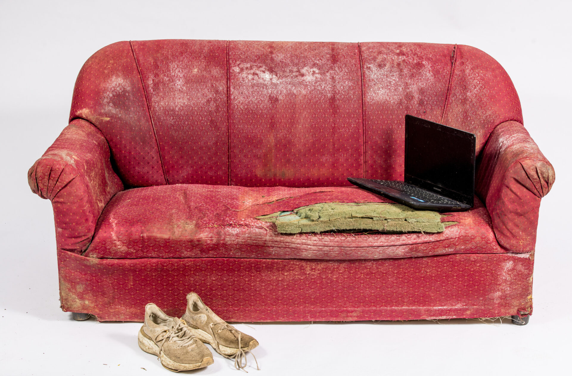 A broken red sofa with stains from floodwaters is photographed with white trainers on the floor in front and an open laptop on the seating area on the right.