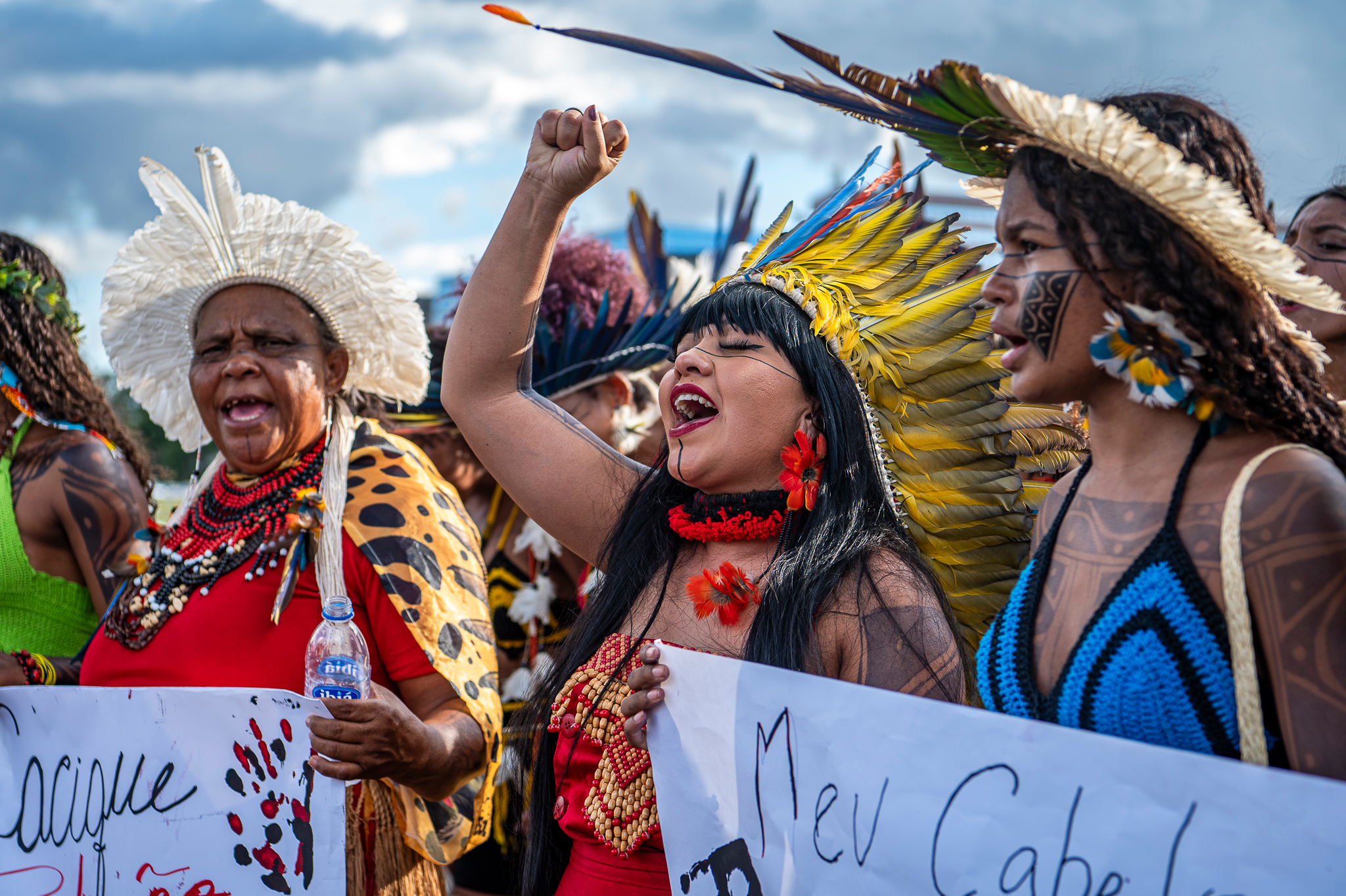 Indigenous women in vibrant traditional dress and feathered headdresses march and chant during a demonstration, holding banners with handwritten messages.