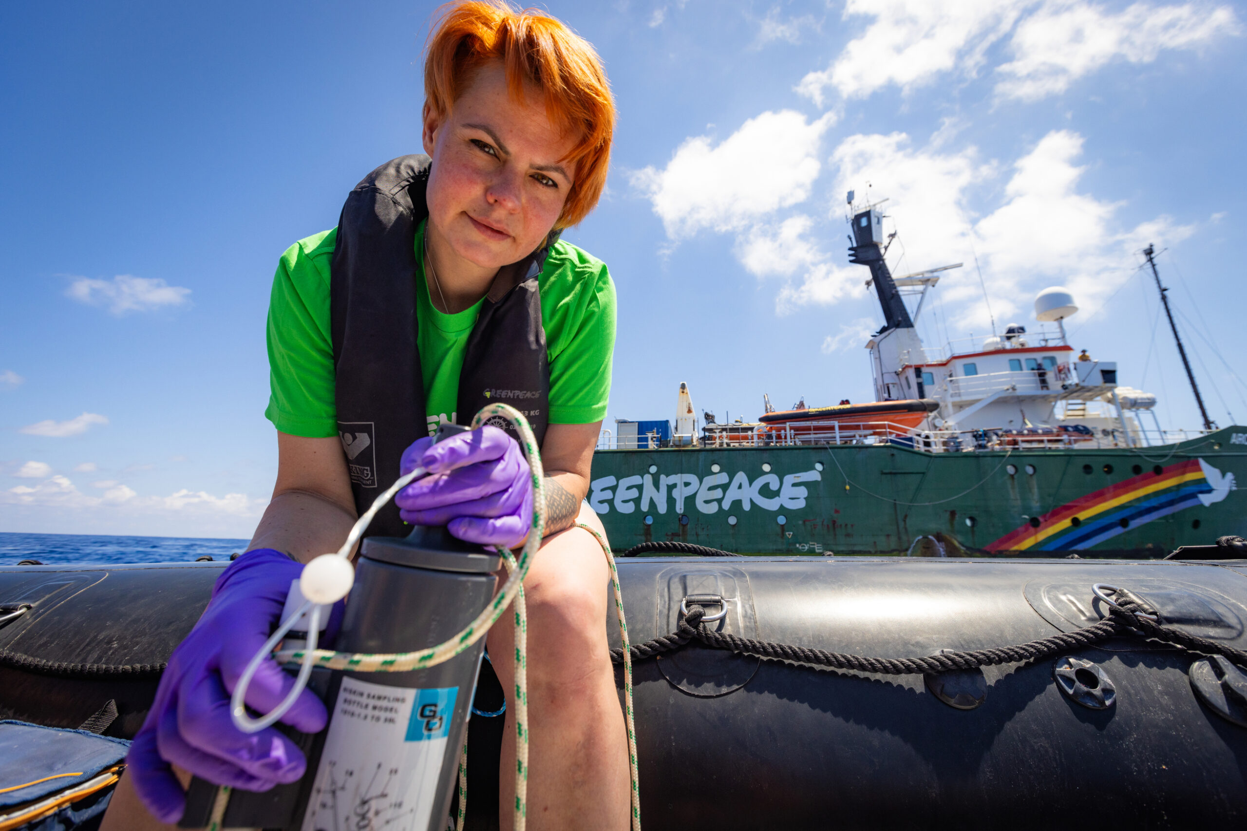 A Greenpeace crew member wearing purple gloves and a life jacket collects a water sample from a small boat, with the Greenpeace ship Arctic Sunrise visible in the background.
