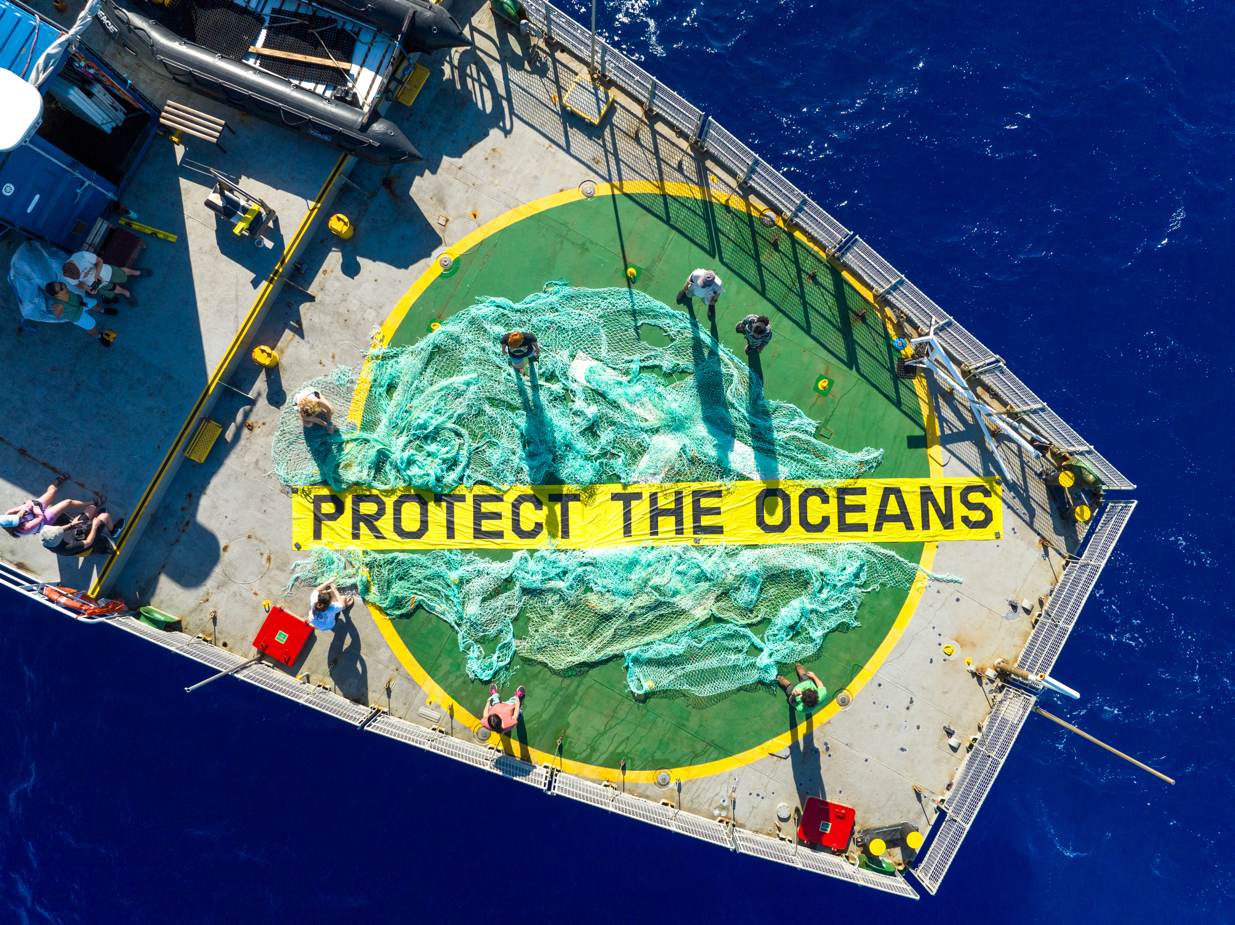An aerial view of the deck of a Greenpeace ship, where activists stand around fishing nets arranged around a large yellow banner reading “Protect the Oceans.”