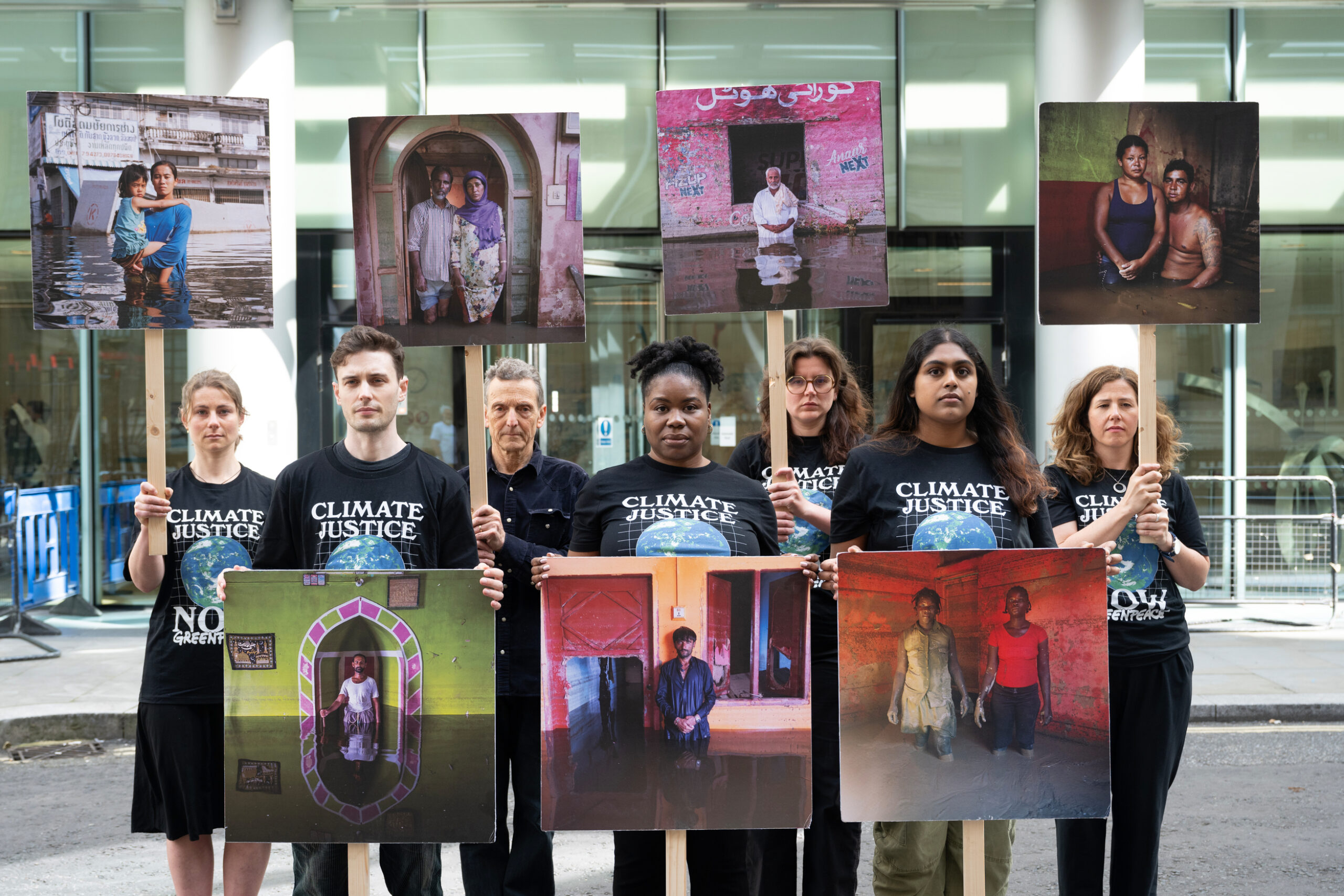A group of climate justice activists stand in a line holding large portraits of people affected by climate change, each image showing individuals in flood-damaged or otherwise compromised homes.