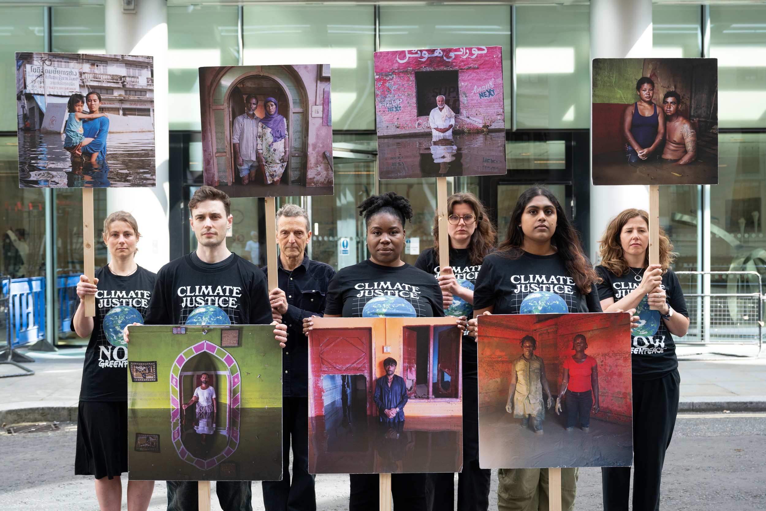 A group of climate justice activists stand in a line holding large portraits of people affected by climate change, each image showing individuals in flood-damaged or otherwise compromised homes.