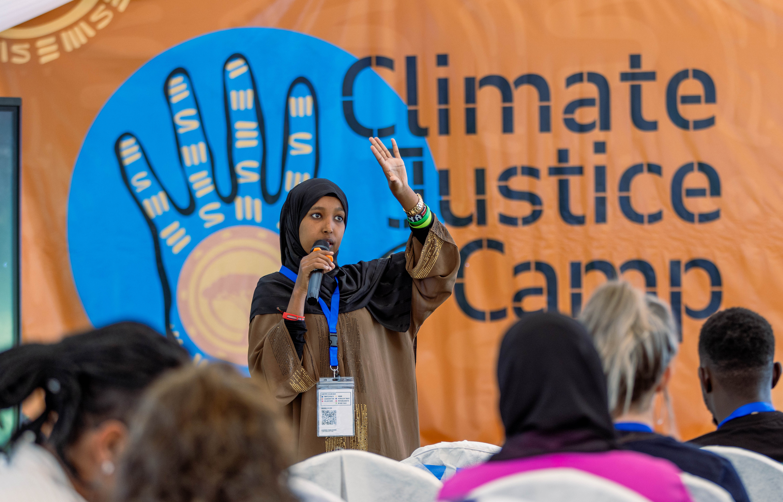 A woman wearing a headscarf speaks into a microphone and gestures during a session at the Climate Justice Camp, with a banner reading “Climate Justice Camp” and a stylised hand symbol behind her.