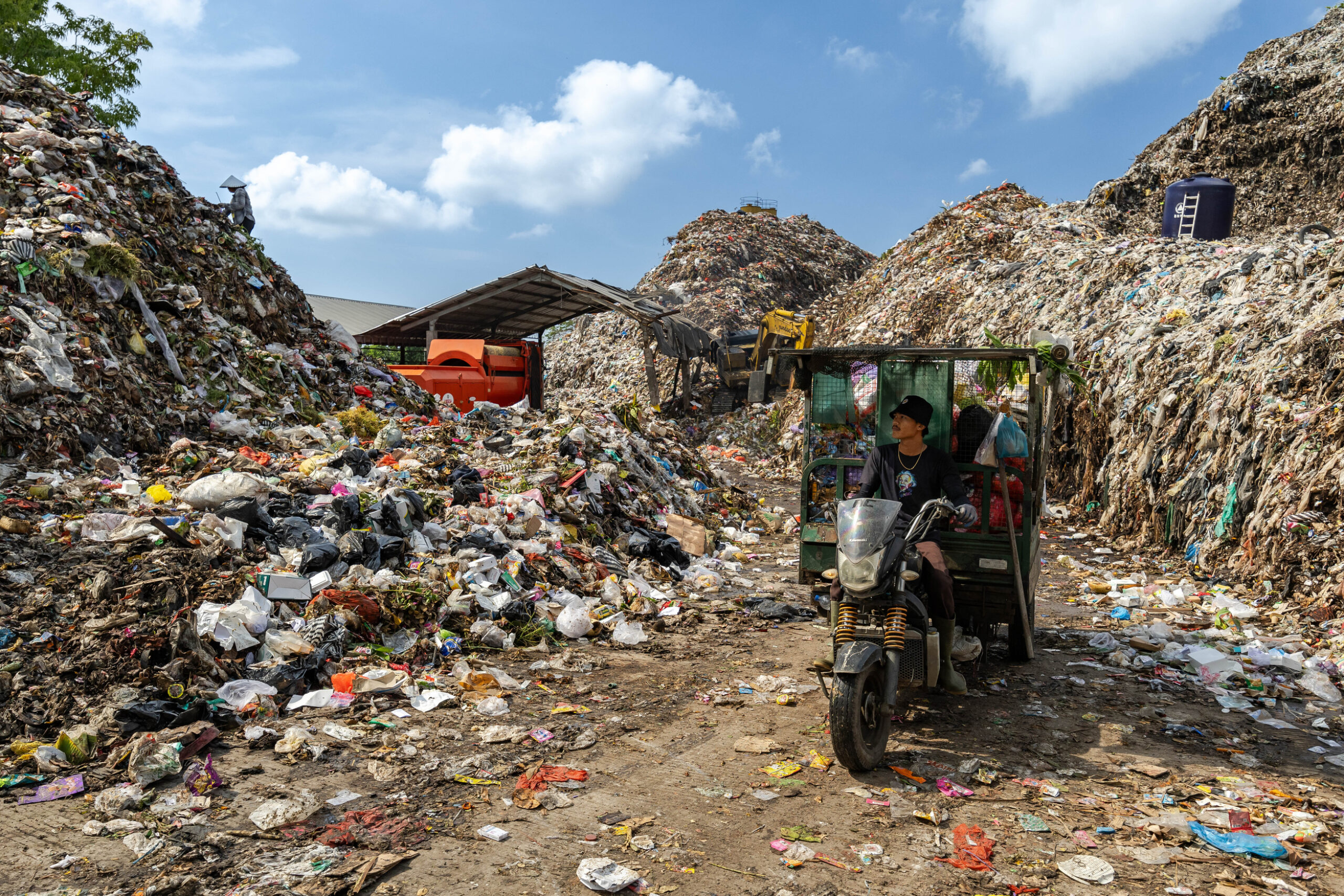 A person drives a three-wheeled motor vehicle through a waste site piled high with plastic and other rubbish, surrounded by towering heaps of garbage under a blue sky with scattered clouds.
