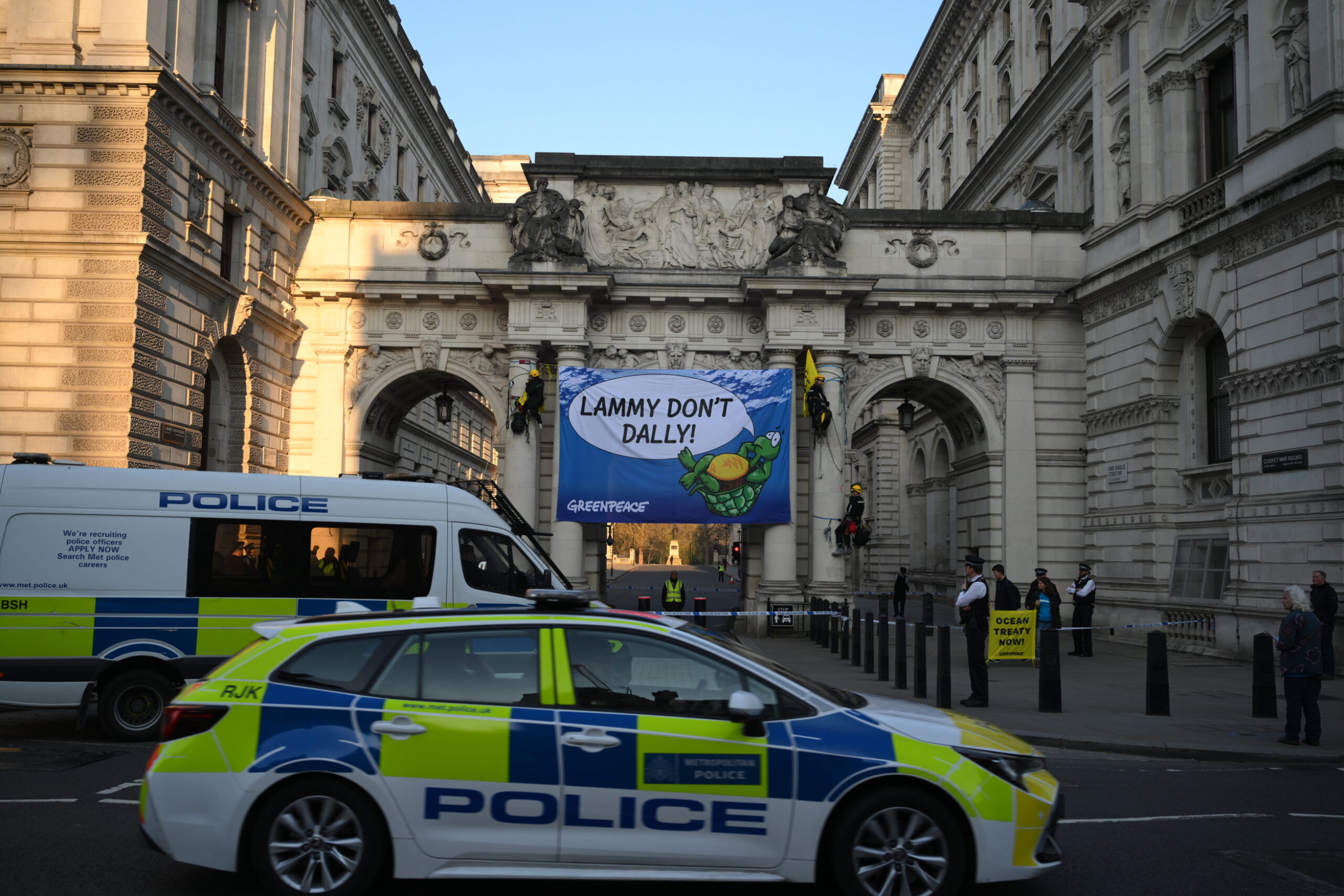 A banner featuring a turtle and the slogan "Lammy Don't Dally!" hangs from the monumental arches at the Foreign Office building in Whitehall, with police cars in the foreground
