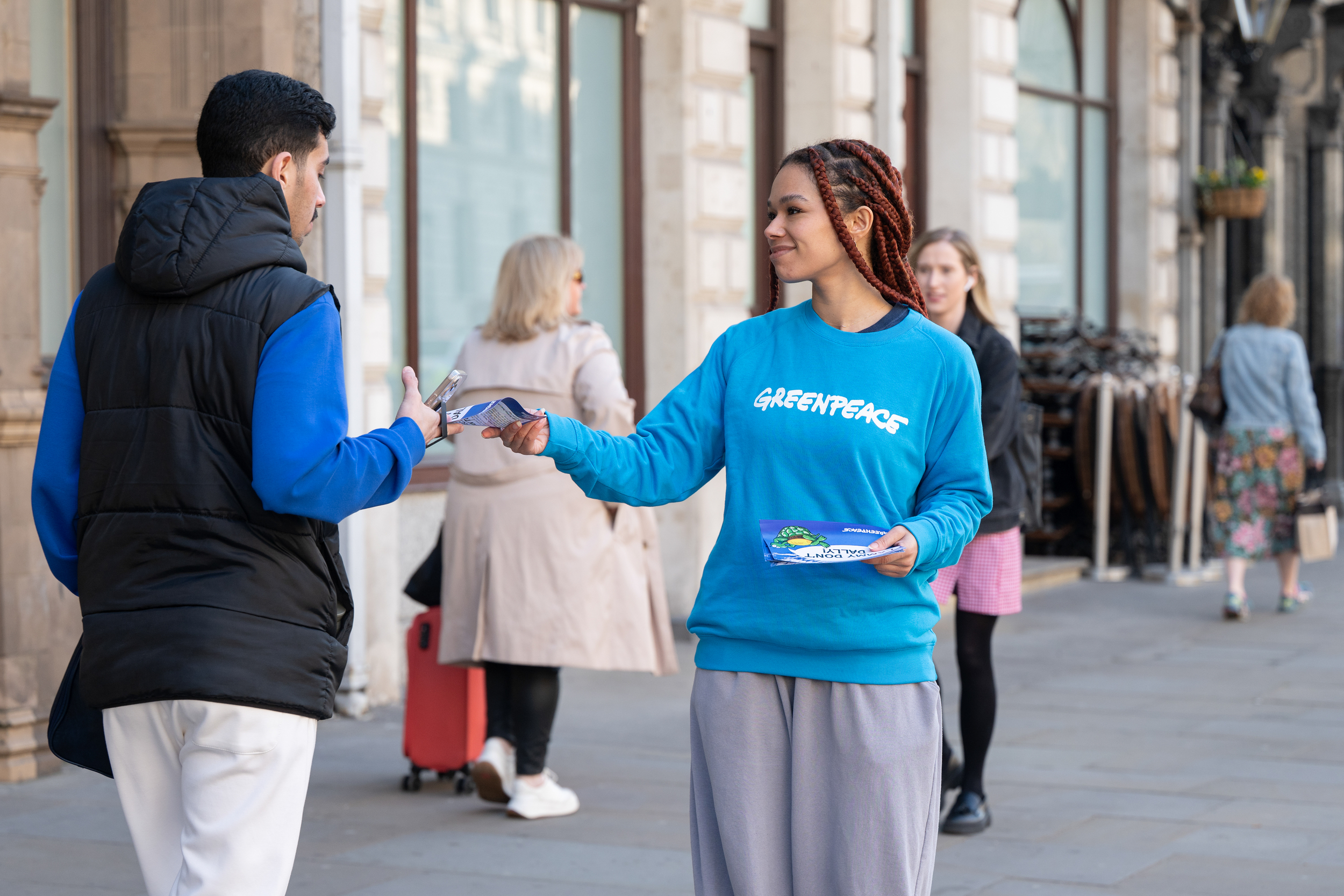 A smiling Greenpeace activist in a blue sweatshirt hands a leaflet to a passer-by on a city street, with other pedestrians walking in the background near a row of stone buildings.