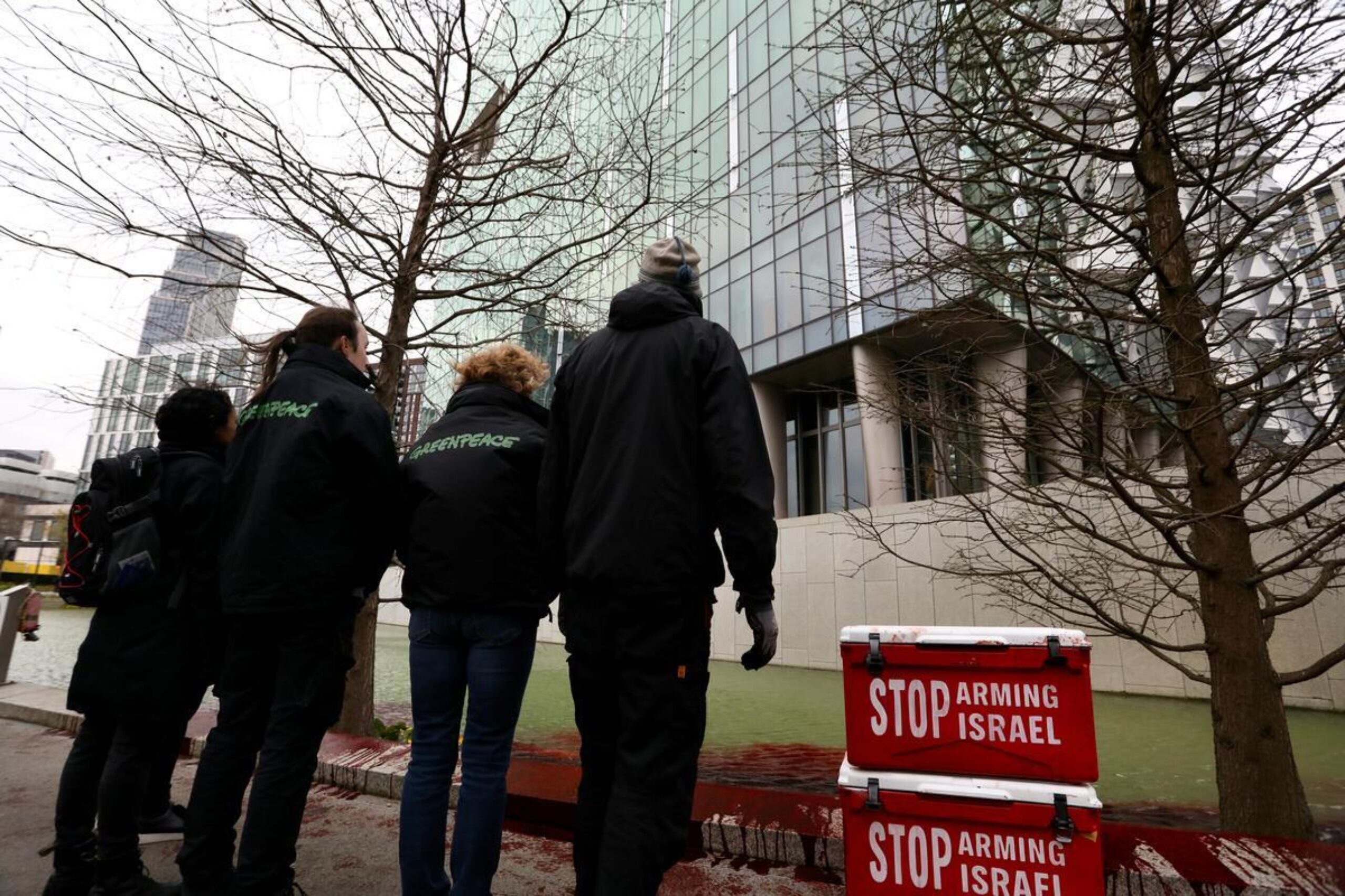 Greenpeace activists look up at the US Embassy building in London. The ornamental pond in front of them is stained with blood-red dye, and there are red containers marked 'Stop arming Israel' stacked beside them.