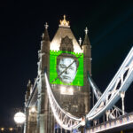 Greenpeace projects a large image of Elon Musk’s face, crossed out with a diagonal black line, onto Tower Bridge in London at night. The background of the projection is bright green with the words “TIME TO RESIST” above the face.