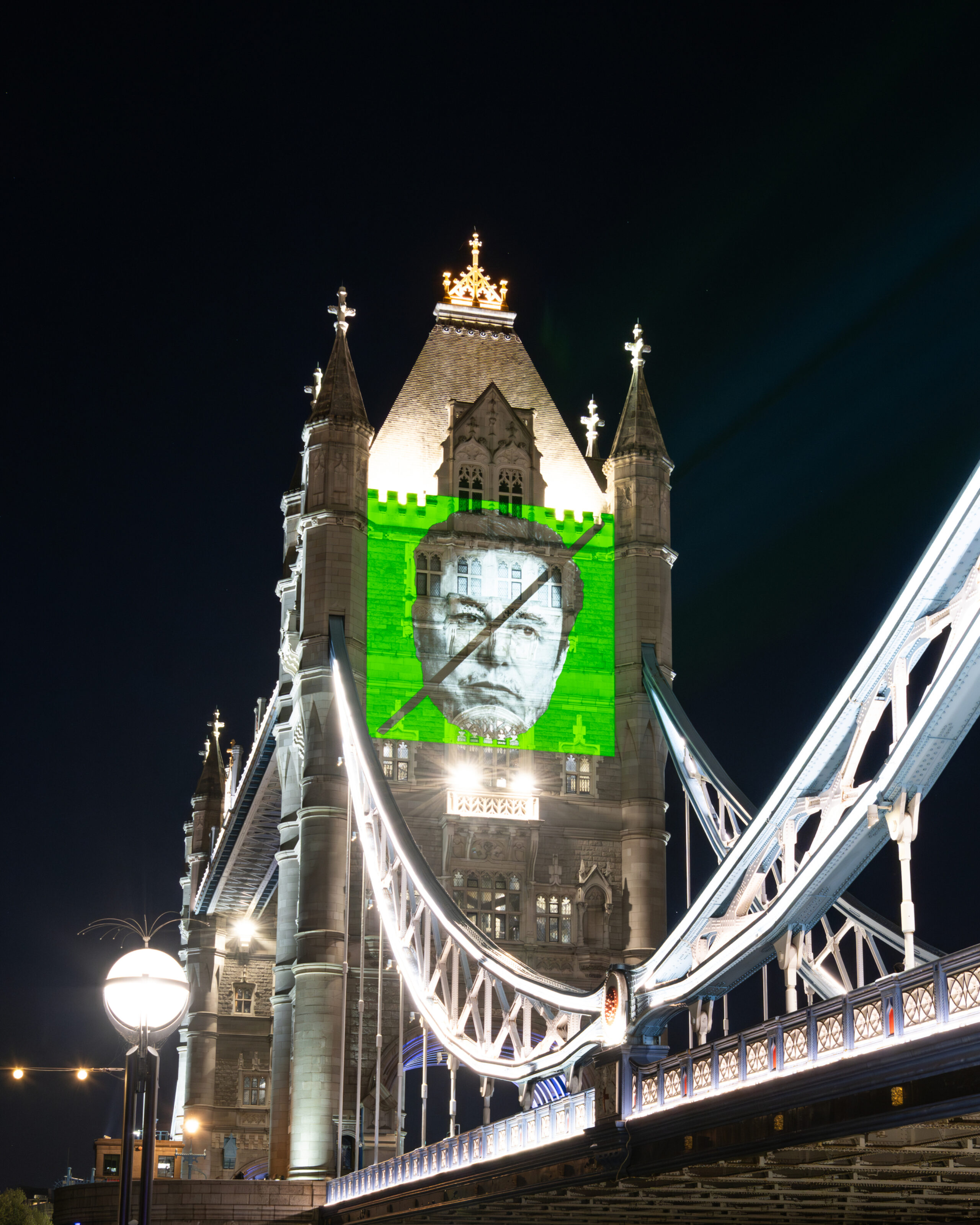 Greenpeace projects a large image of Elon Musk’s face, crossed out with a diagonal black line, onto Tower Bridge in London at night. The background of the projection is bright green with the words “TIME TO RESIST” above the face.