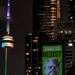 Greenpeace projects an image of Donald Trump’s face, crossed out with a black diagonal line, and the words “TIME TO RESIST” in bold green lettering onto a high-rise building in Toronto at night. The CN Tower, lit in rainbow colours, is visible in the background.