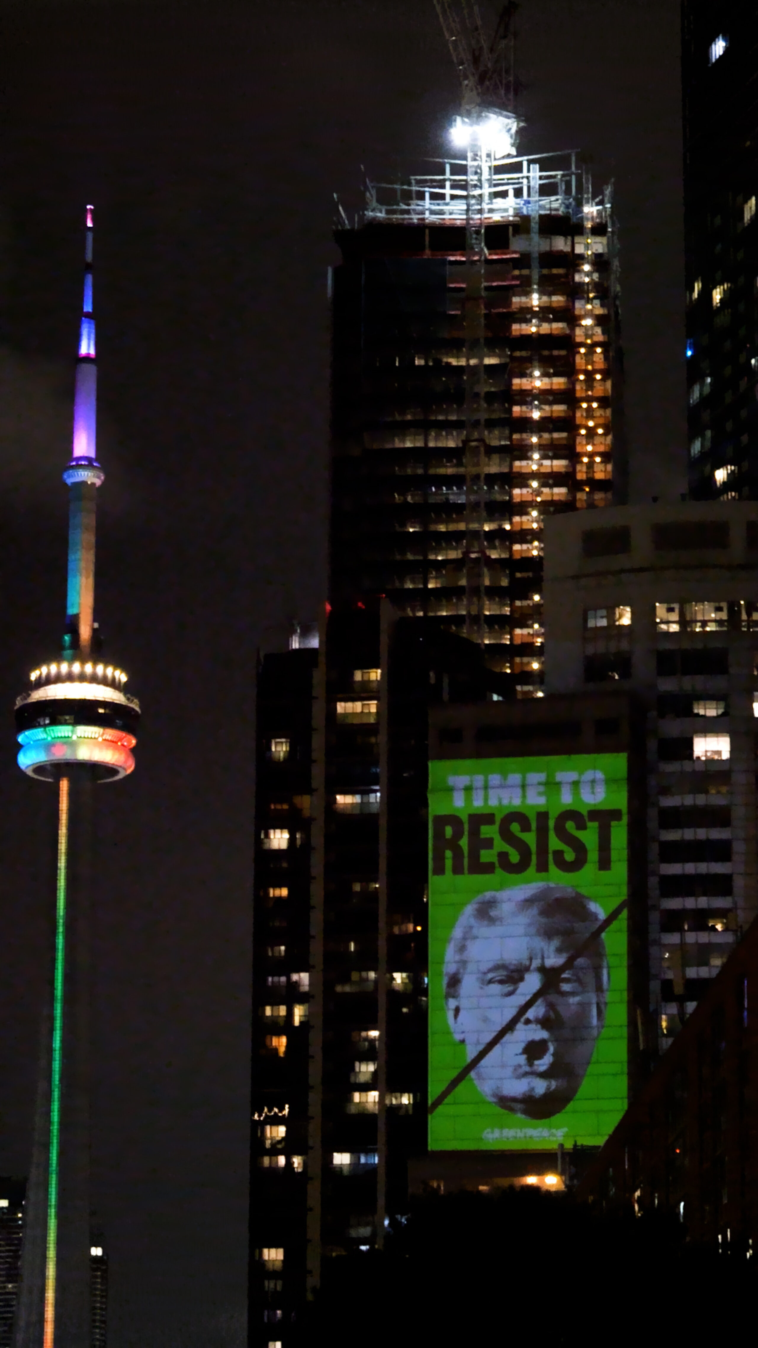 Greenpeace projects an image of Donald Trump’s face, crossed out with a black diagonal line, and the words “TIME TO RESIST” in bold green lettering onto a high-rise building in Toronto at night. The CN Tower, lit in rainbow colours, is visible in the background.