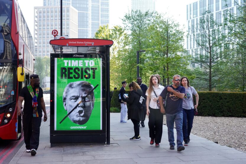 A Greenpeace billboard at a London bus stop near the US Embassy displays a green background with the face of Donald Trump, crossed out with a black line, and the words “TIME TO RESIST”. Passers-by walk past the scene in an urban setting.