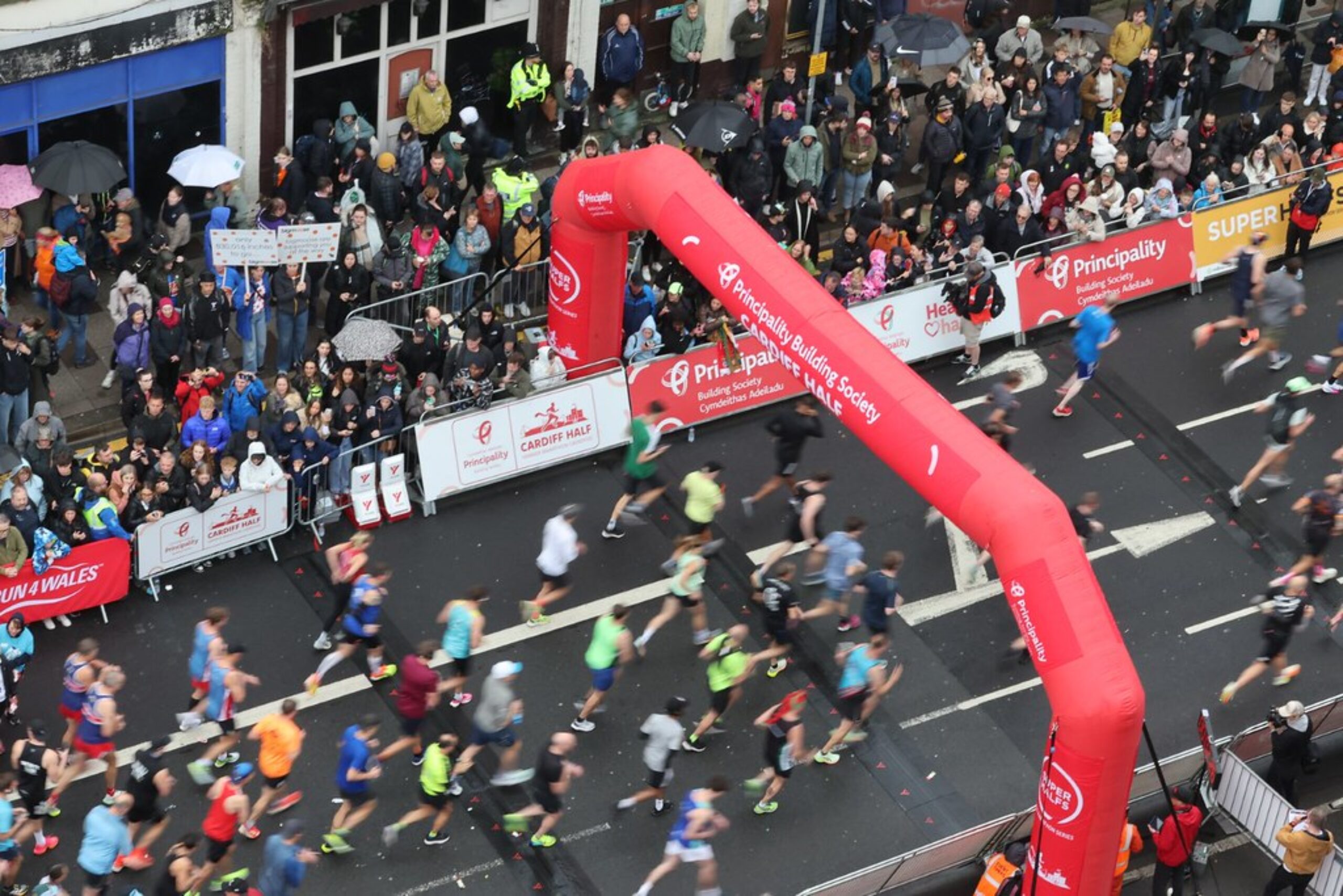 Aerial view of runners crossing under a red inflatable arch in the Cardiff Half Marathon, with crowds cheering behind barriers along the wet city streets.