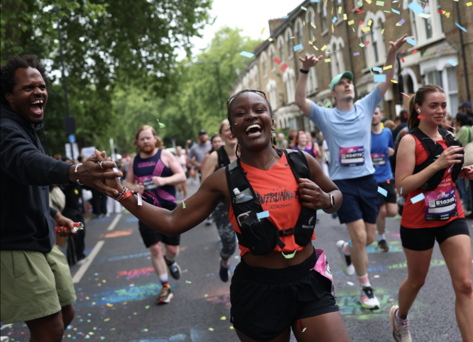 A joyful runner high-fives a spectator while confetti falls around her in a bustling city street race, surrounded by cheering crowds and fellow runners.
