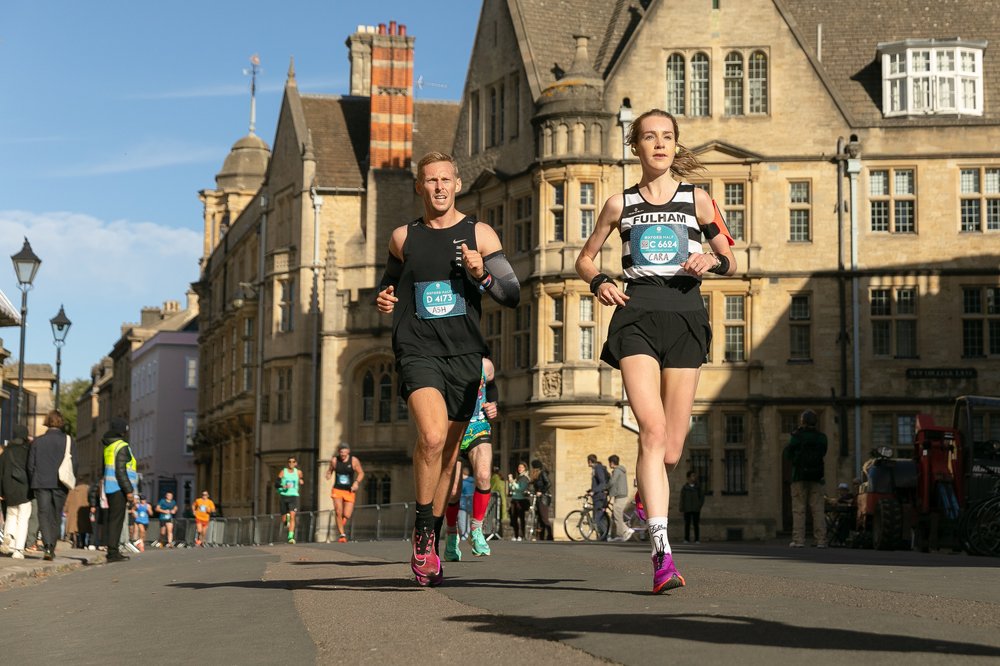 Two runners from Fulham Running Club approach the camera during the Oxford Half Marathon, with Oxford’s historic architecture visible behind them.
