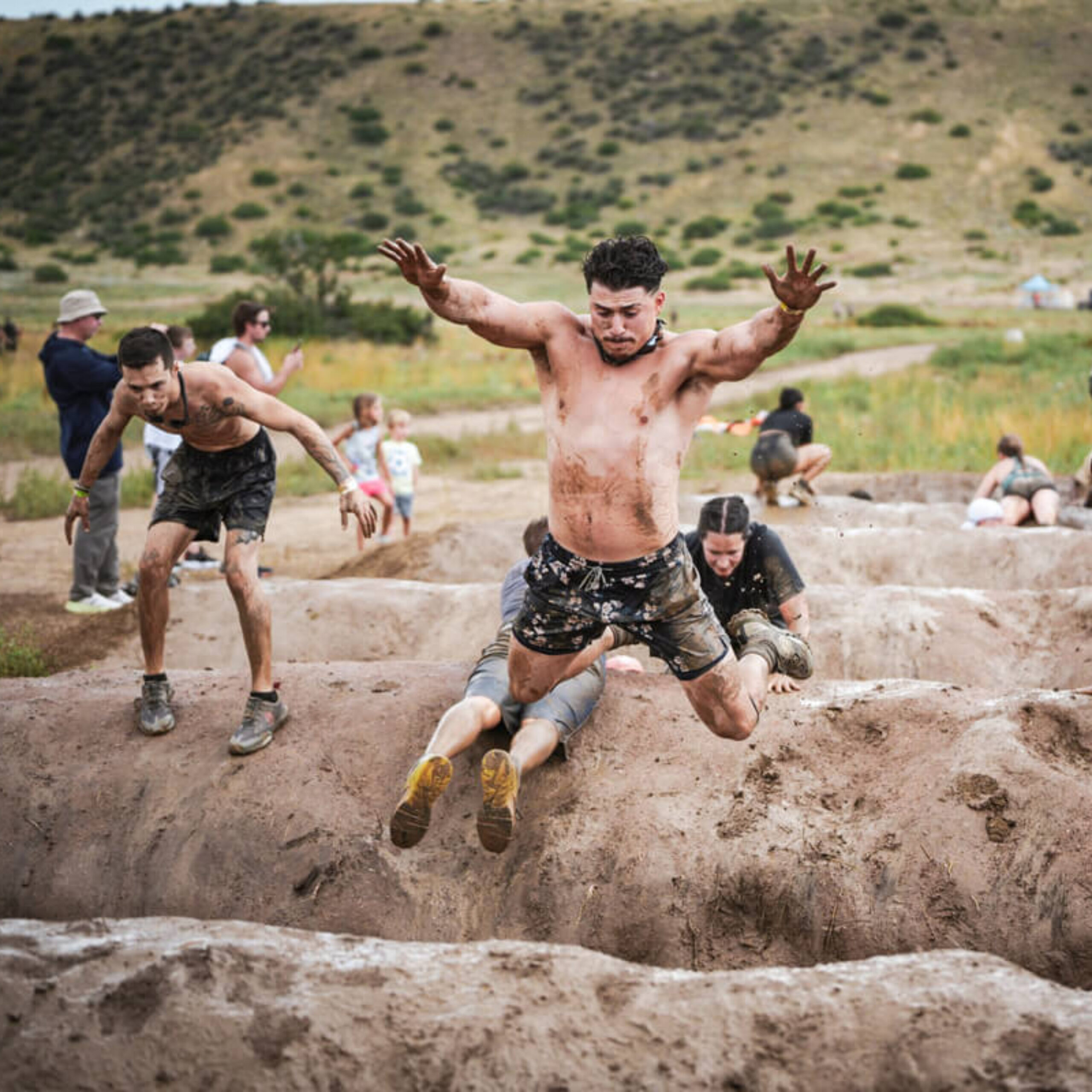 A shirtless man jumps into a muddy obstacle while others climb and crawl through trenches during a Tough Mudder endurance challenge.