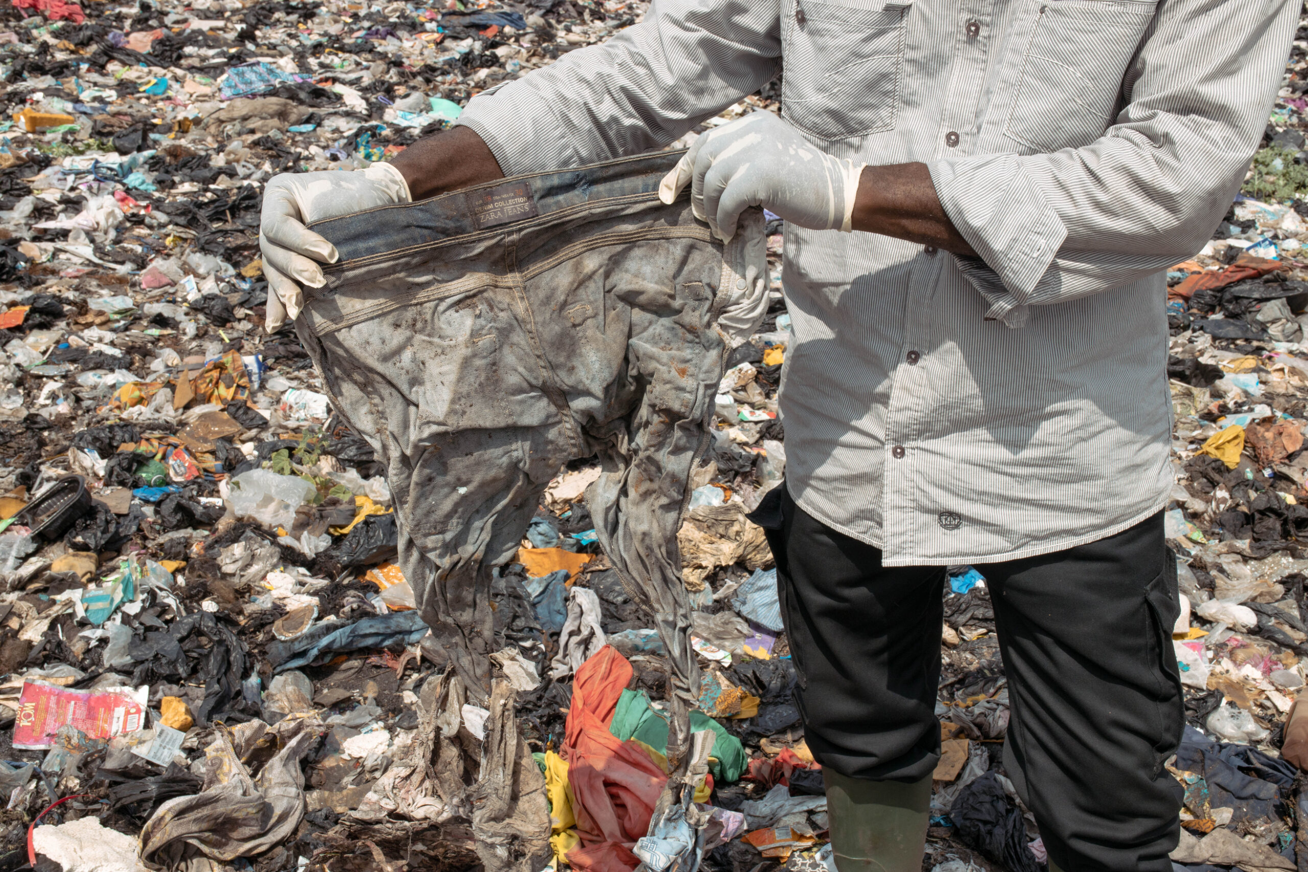 A man wearing gloves holds up a pair of muddy, damaged jeans labelled “ZARA JEANS” in the middle of a massive textile and plastic waste dump.