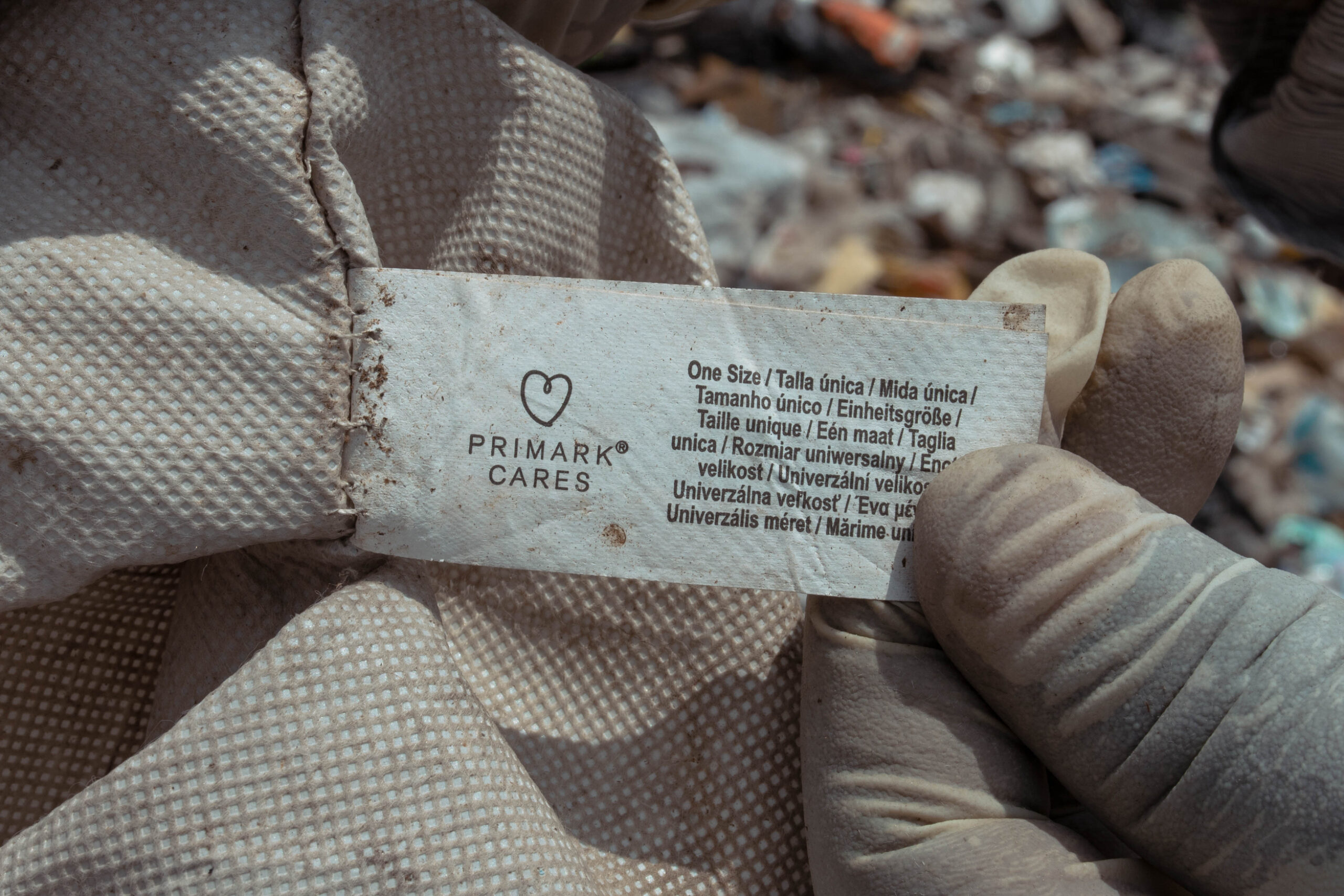 Close-up of gloved hands holding a dirty garment label that reads “Primark Cares”, found among textile waste in a landfill.
