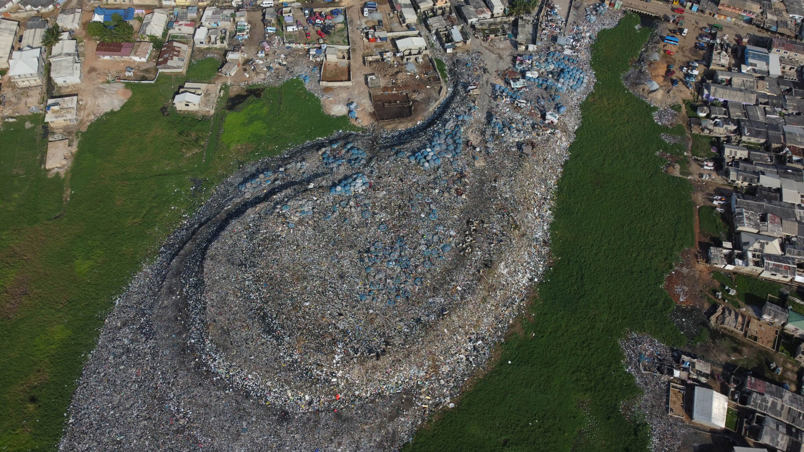 High-angle aerial image of a sprawling landfill site surrounded by green areas and dense urban housing. The shape of the site makes it seem like the waste is flowing towards the green space like liquid.