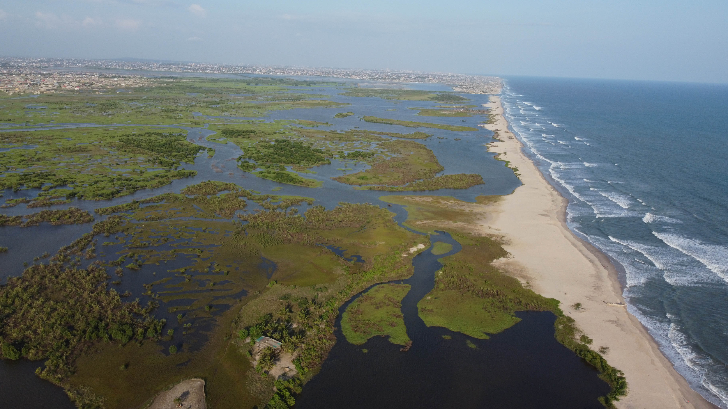 Drone shot of a green, marshy wetland area meeting a long, sandy coastline with ocean waves on one side and a densely populated cityscape on the horizon.