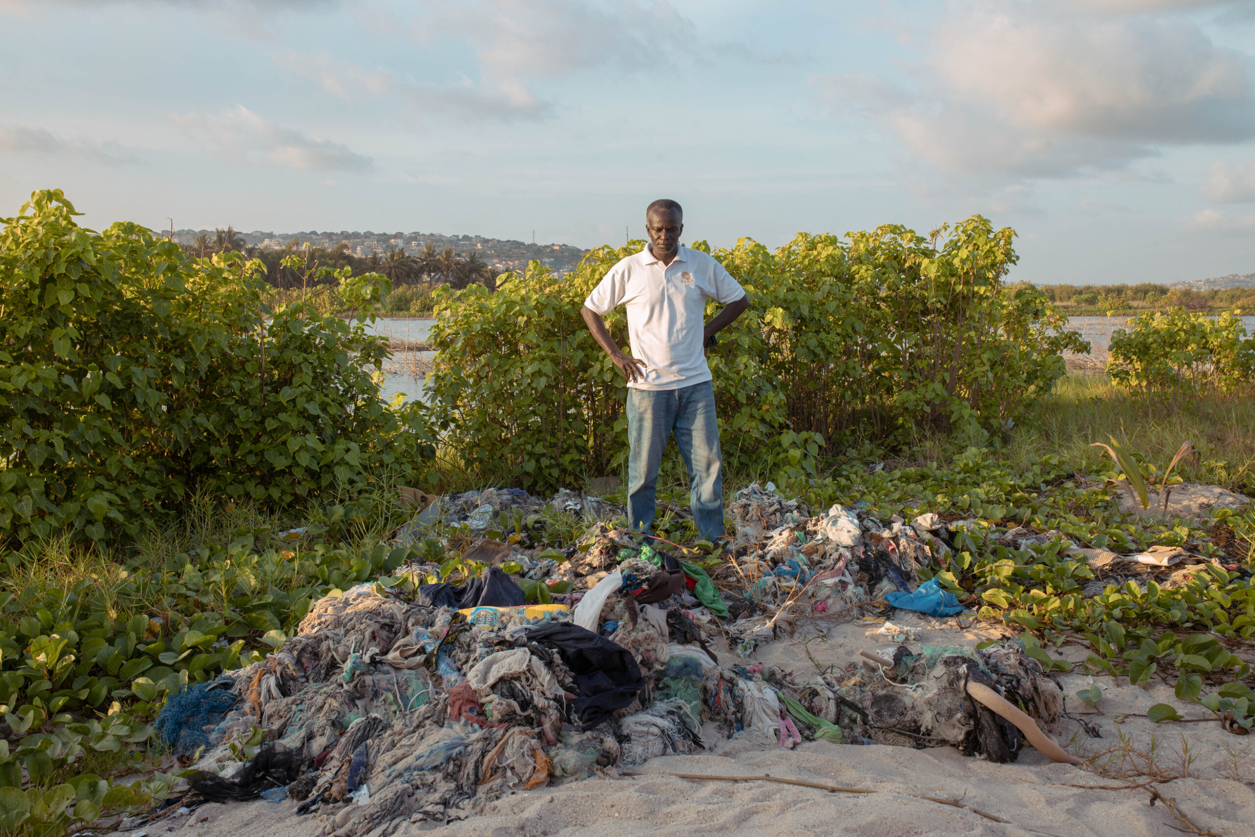 A man stands with hands on hips next to a mound of textile waste scattered across a sandy area with dense green vegetation and a river behind him, in a semi-rural environment.