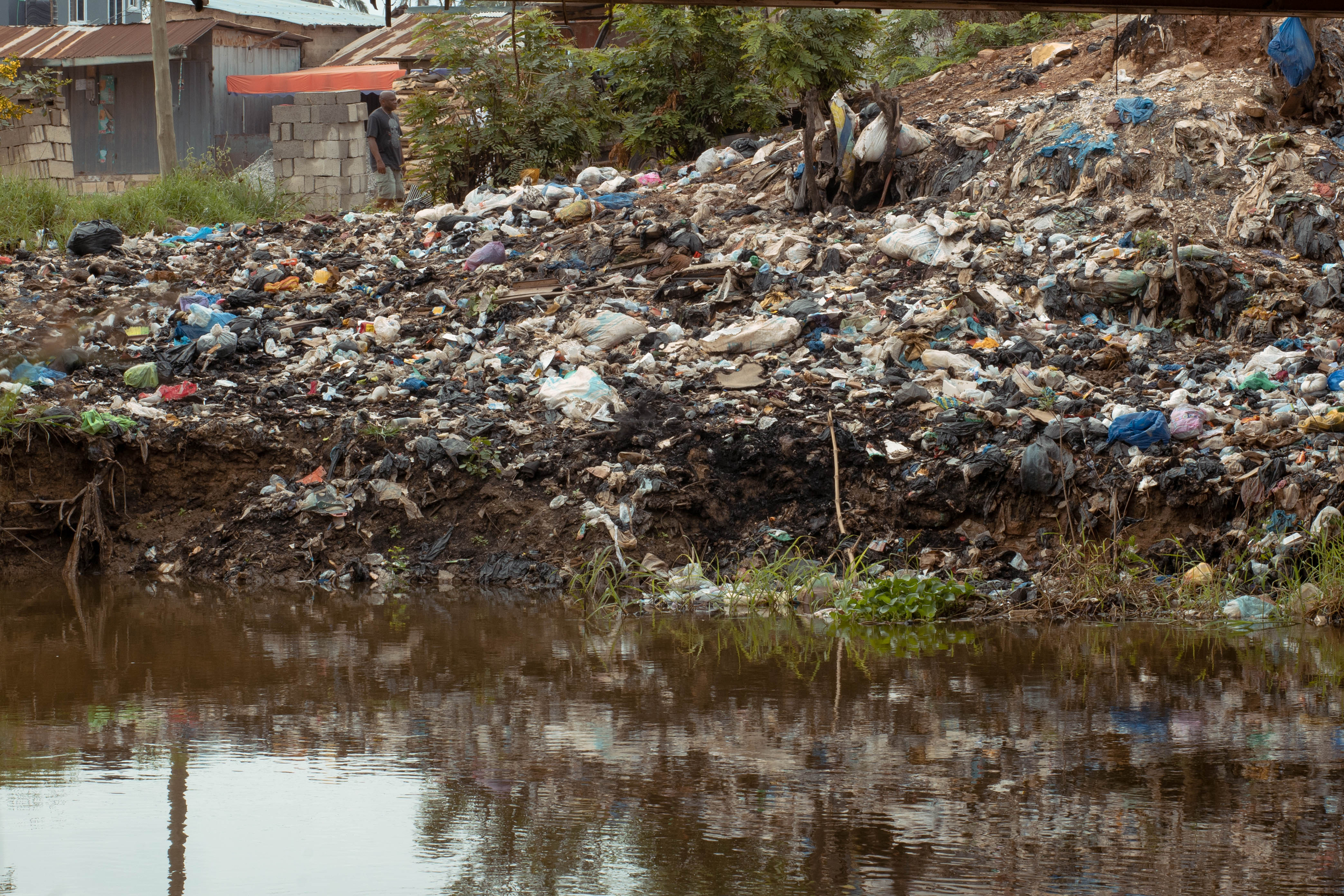 Large heap of plastic, fabric, and general waste piled on the bank of a brown, stagnant water body, with nearby buildings and vegetation visible in the background.
