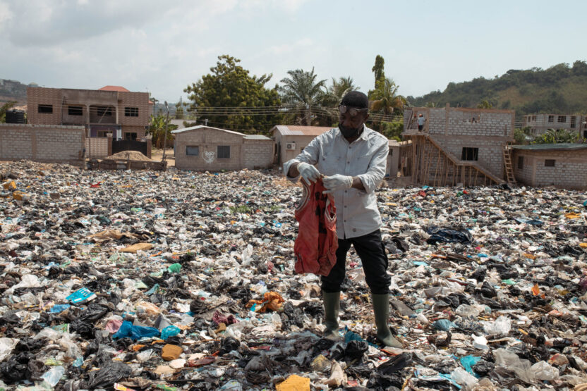 A man wearing gloves and boots examines a red garment while standing in a vast landfill covered in discarded clothing and plastic waste, surrounded by partially built homes in a residential area.