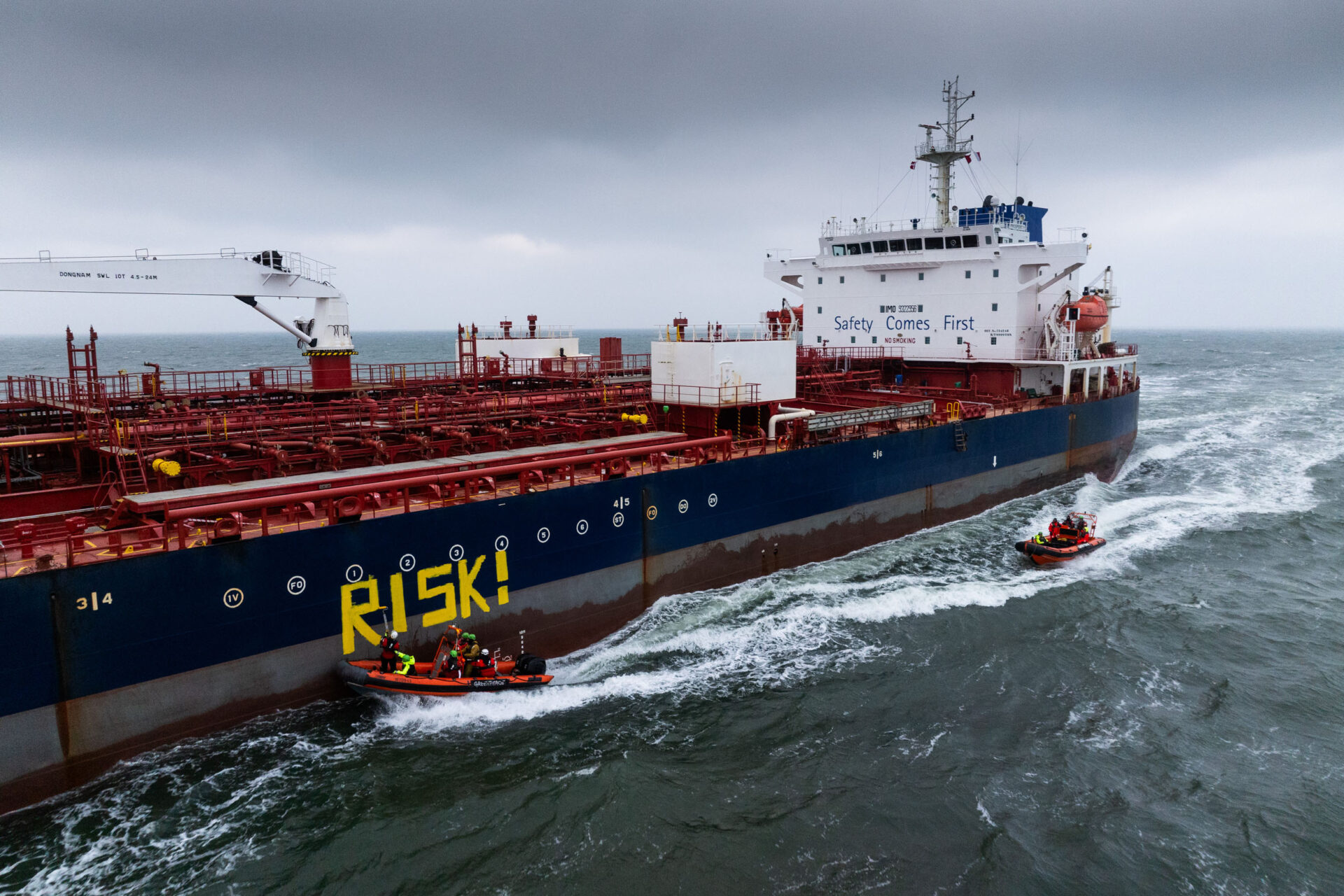 Two inflatable boats drive next to a large tanker and paint "Risk" on the side.